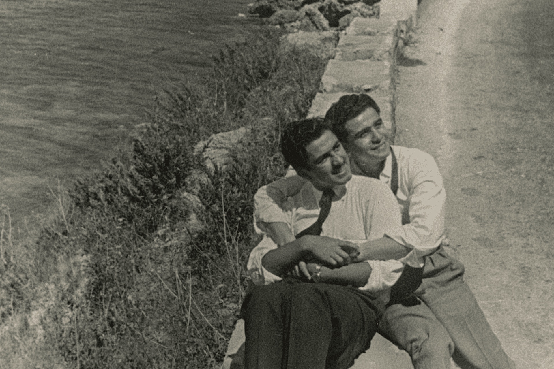 Two figures sit on a stone boundary between the shore and the sea. They embrace one another. 