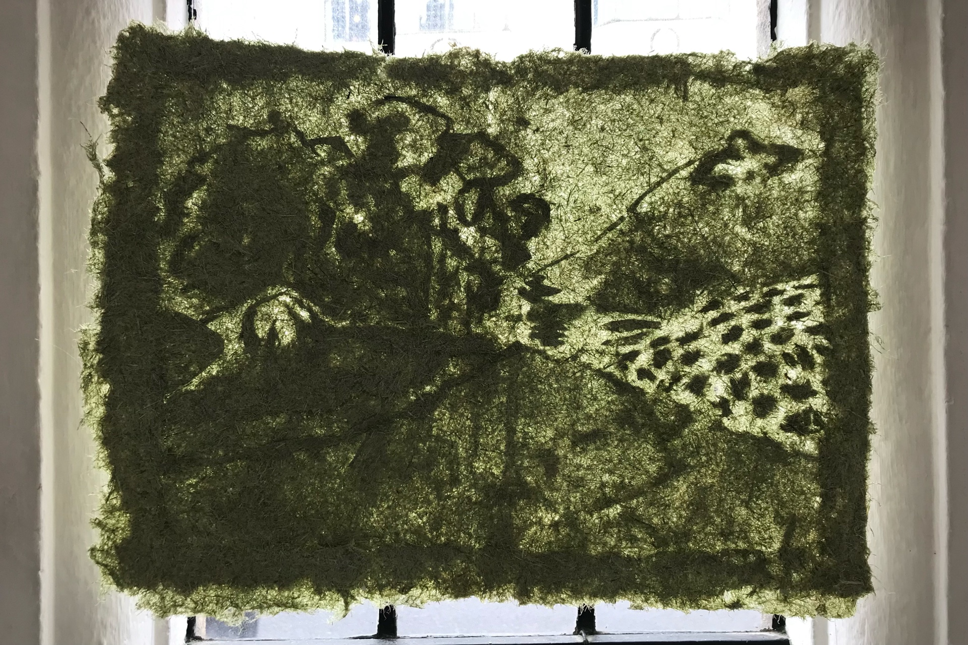 A grass piece of paper hanging in a in front of window, the light coming through the grass paper rending a landscape depicting two hill peaks that are breasts