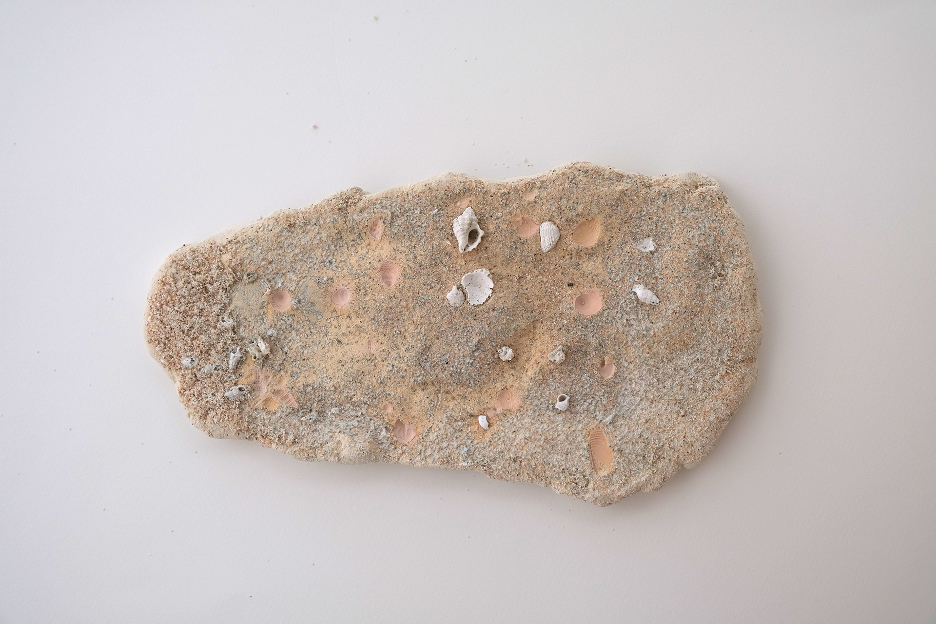 An irregular slab of clay with sand and shells pressed on it. The piece is fired together after the impression was made causing the sand and shells to react to the heat. The shells turned white and the silica turned blue.