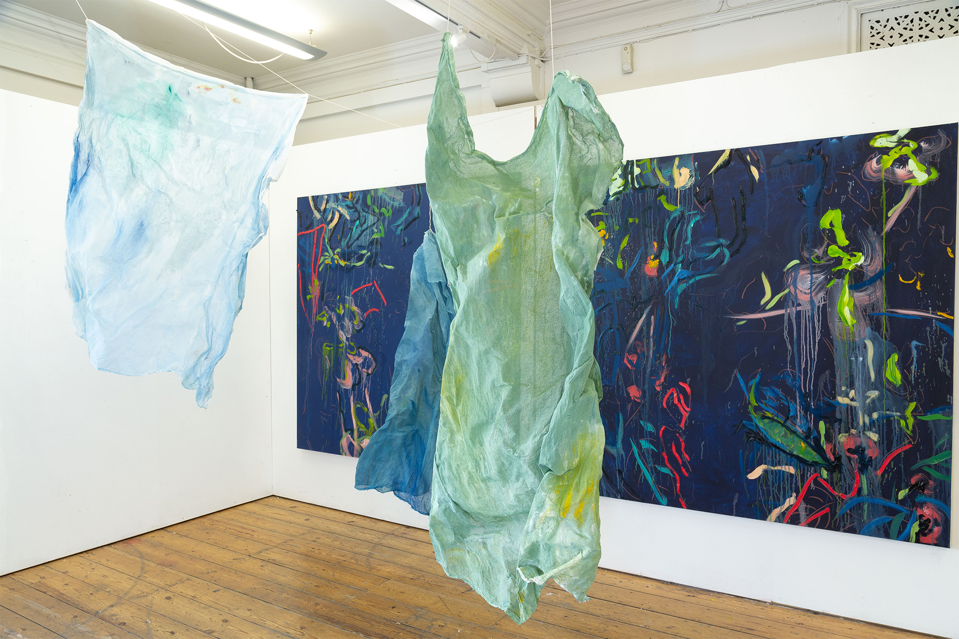 Green and blue fabric hanging infront of two blue paintings each 180cm by 180cm, colourful and gestural brush strokes.