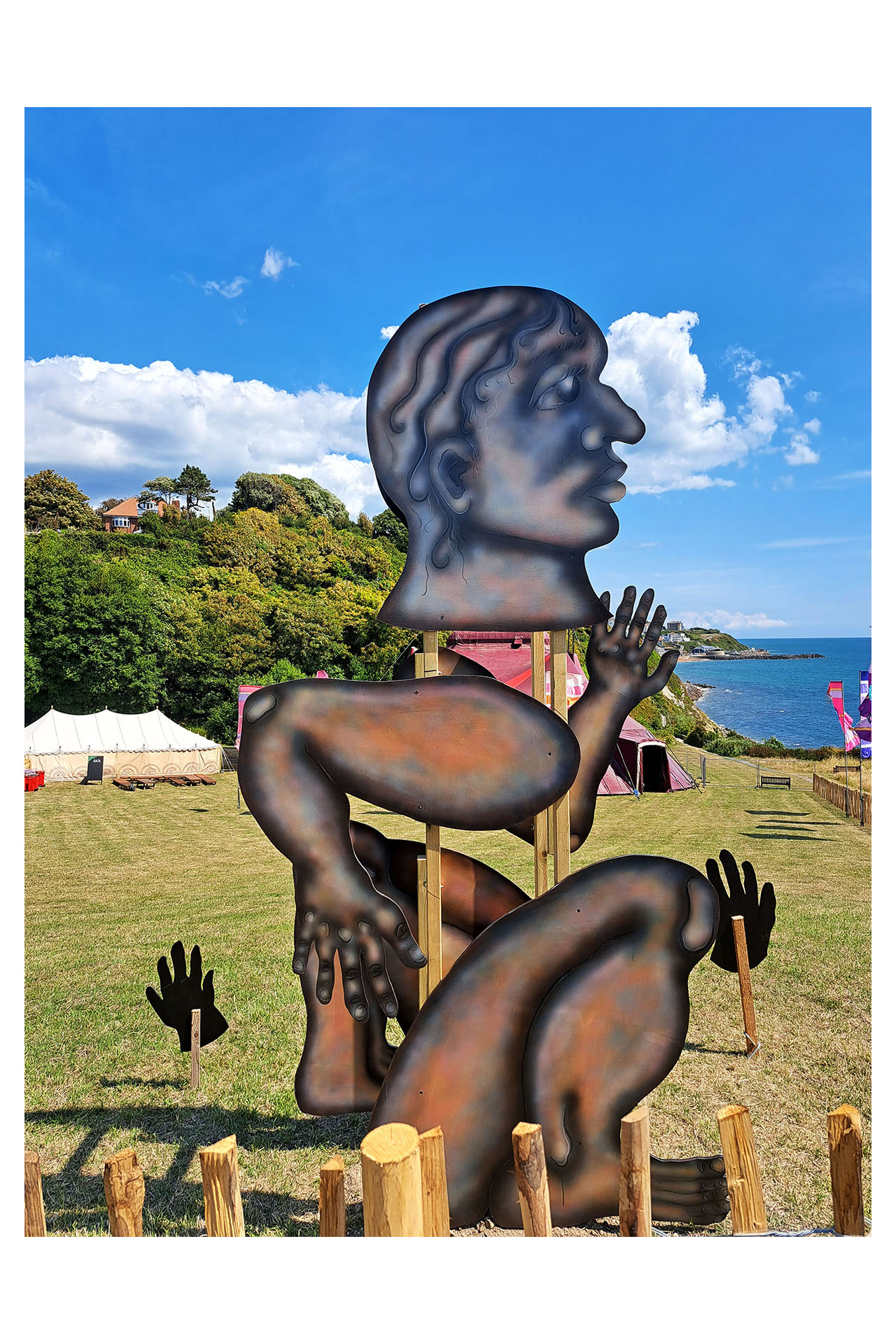 A 250cm tall figure constructed of tall wooden beams golding limb armatures. It is airbrushed with acrylics, and shows a person with knelt on the ground, ready in position as though about to run. It is displayed outside in a festival backdrop.