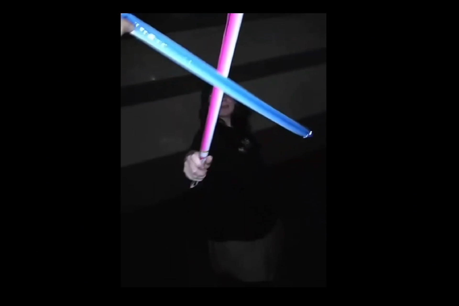 A video still of a woman holding an inflatable neon pink lightsaber, in a dark vignette flash, with another neon blue lightsaber crossing over from another hand, as they swordfight. 