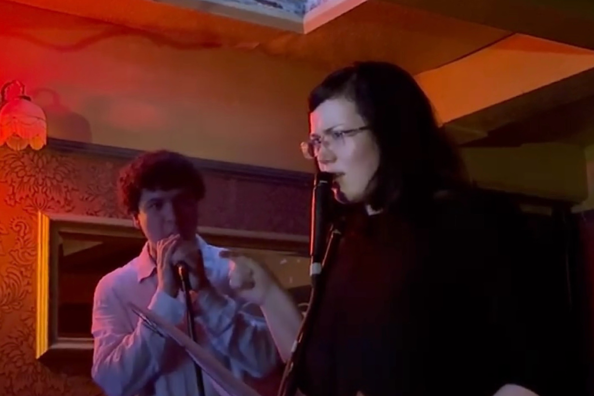 Two figures perform with microphones, within orange stage lighting. 