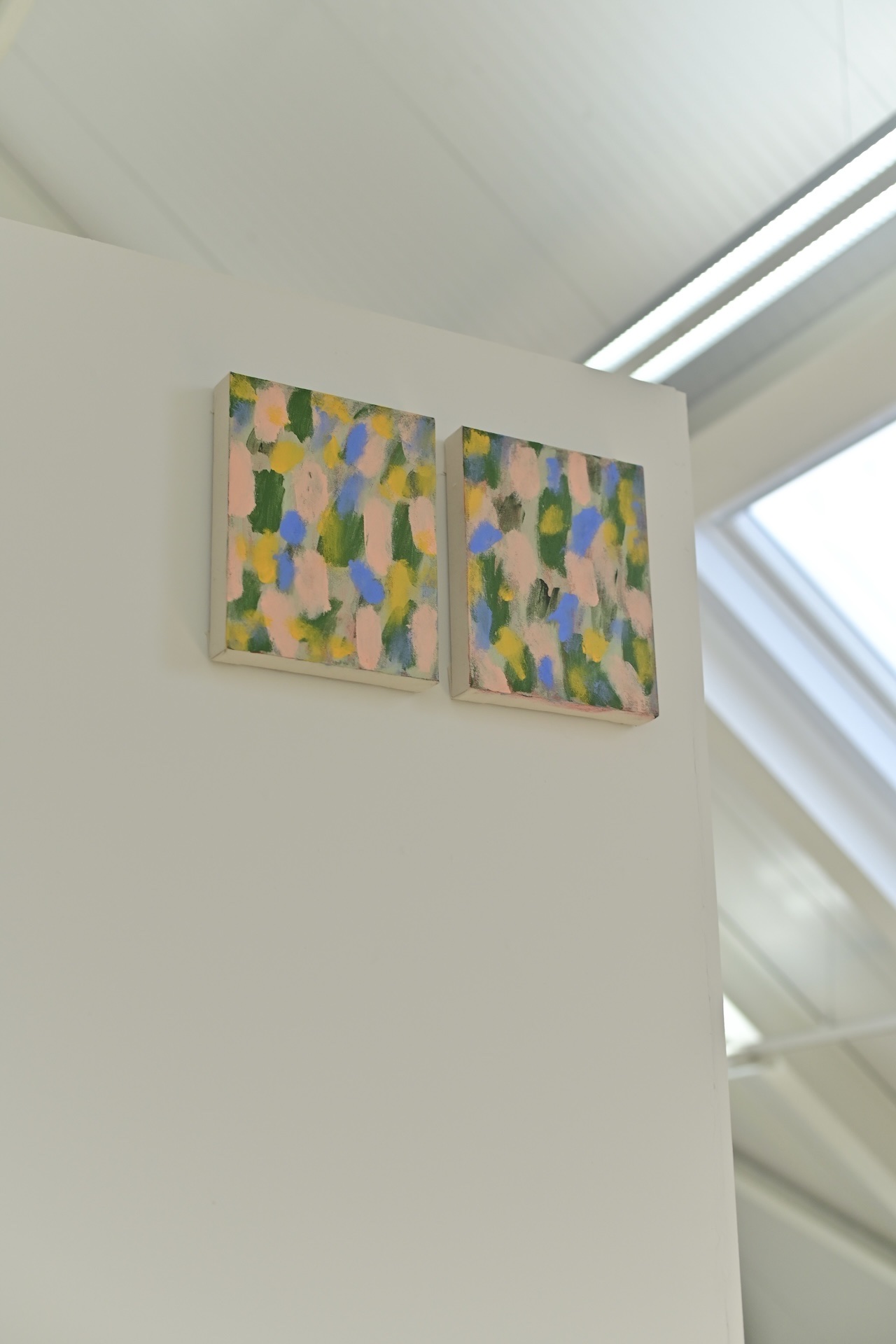Two paintings with blue, green and pink hues on a wooden wall