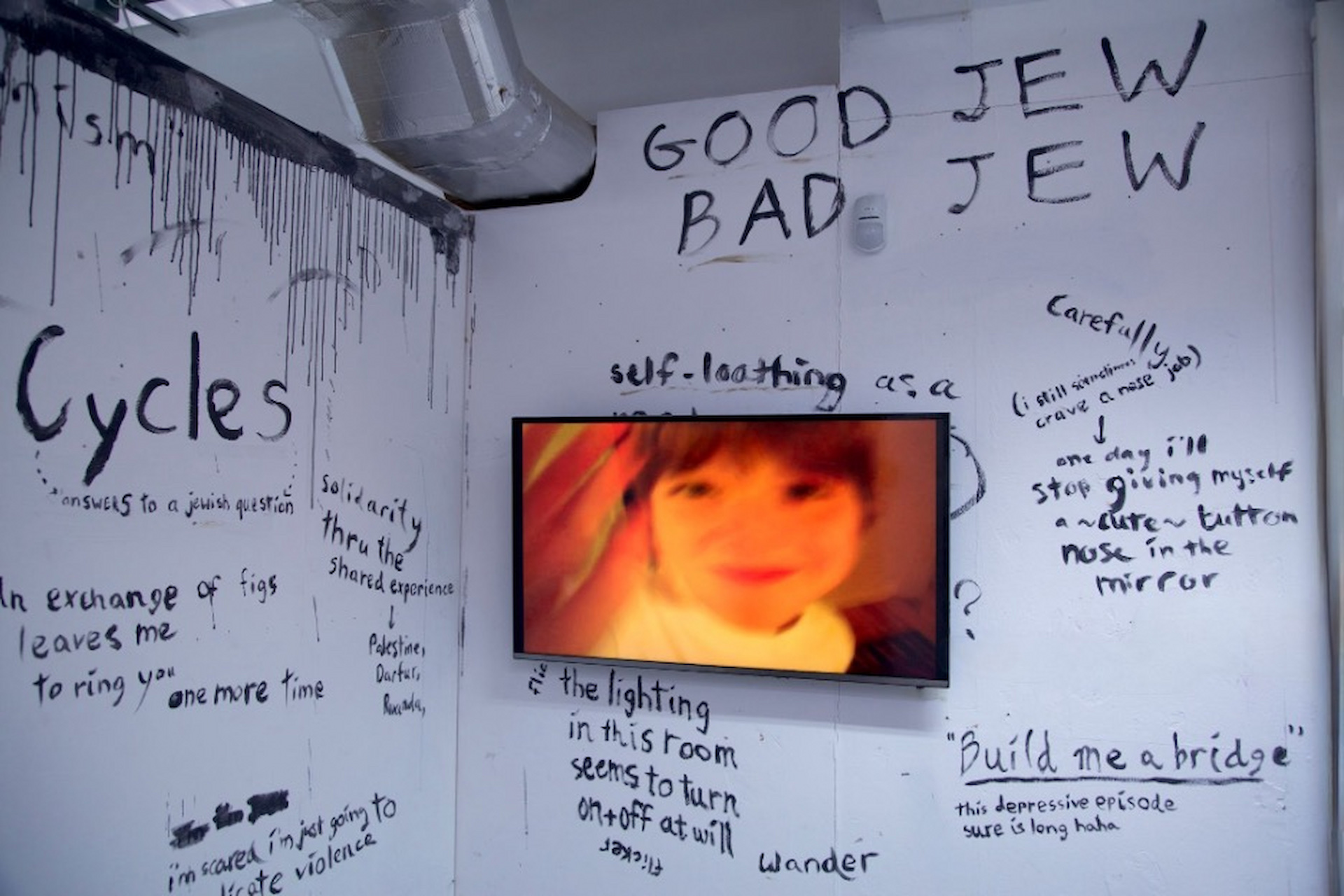 A TV showing childhood archival footage, mounted on a wall adorned by scrawled text in acrylic paint.
