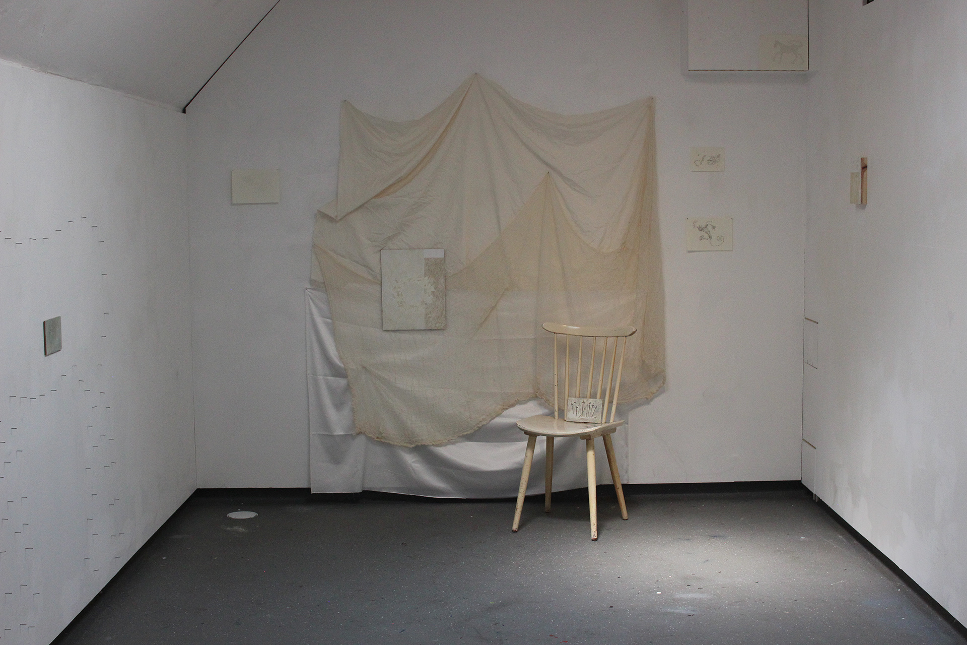 An installation shot of a space with three walls visible. Left wall is cropped but shows a fragment of a work that consisted of small oil paintings and nails. Centre wall is partially draped in  cream coloured fabric, has 4 small scale drawings on paper, 2