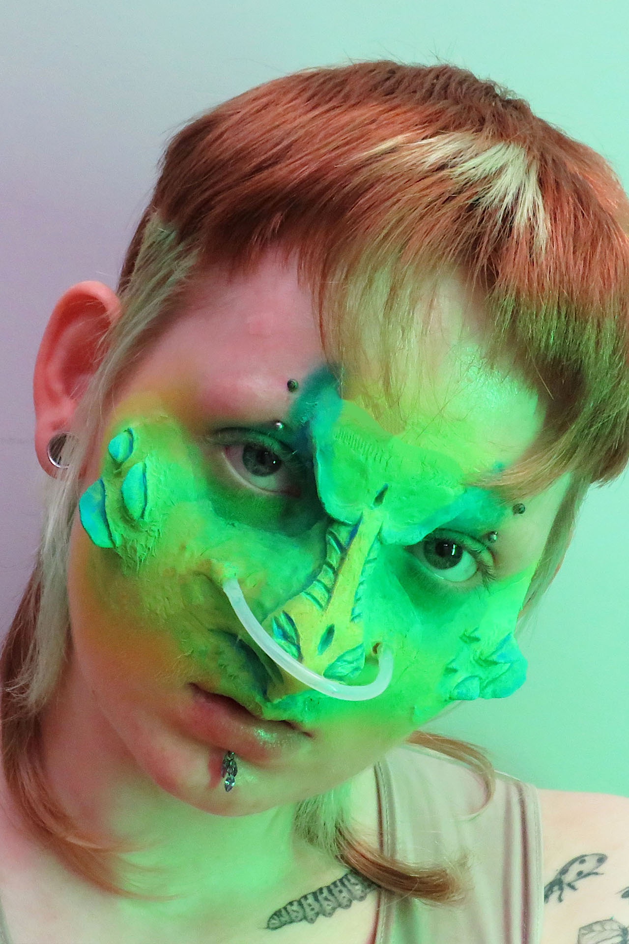 The image shows human-like figure staring directly at the camera. They appear mostly human but have alien-like facial features. On the cheekbones, they have blue and neon green scales protruding, which merge into the almost reptilian nose, which is neon gr