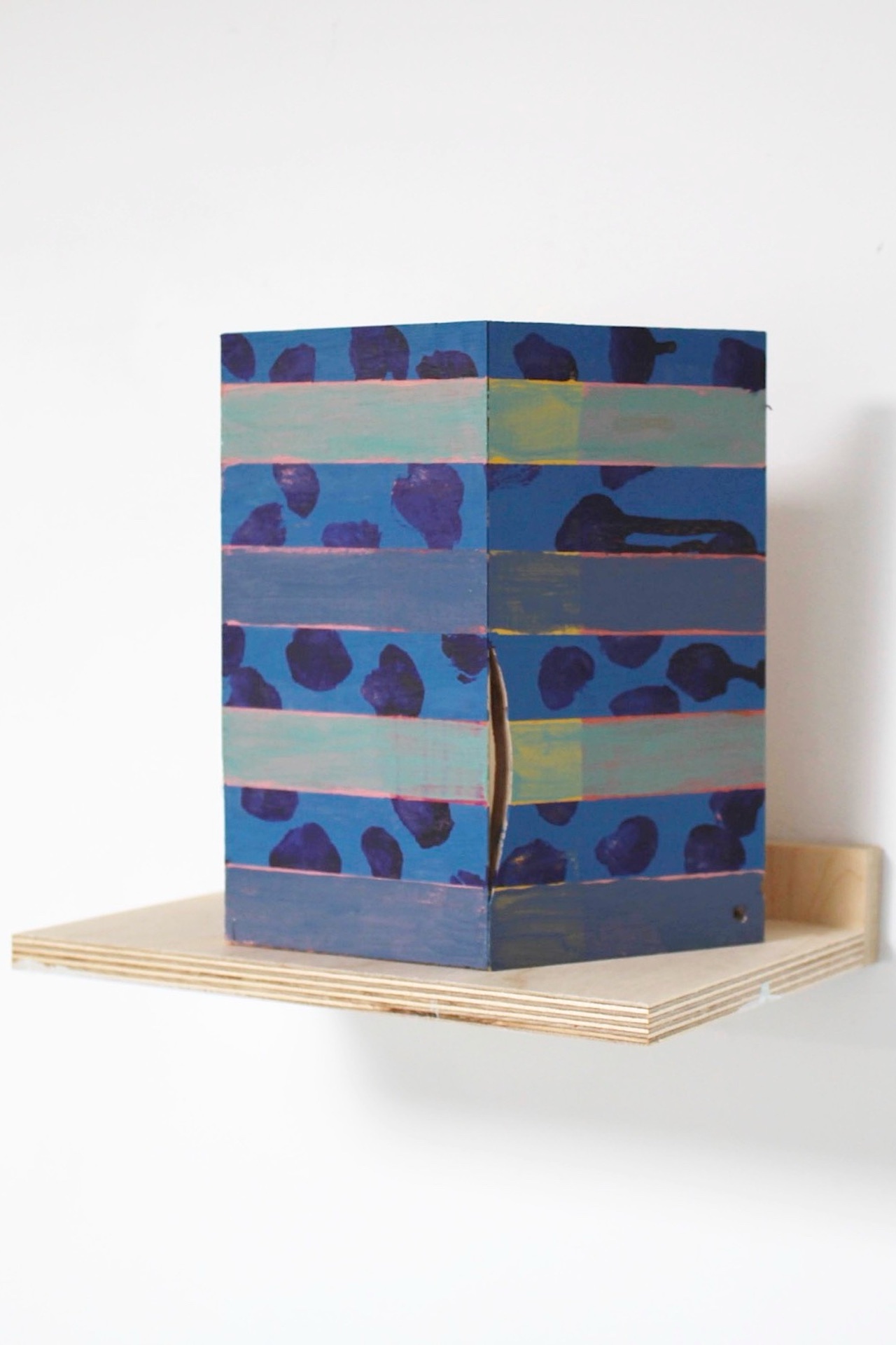Side profile of a painted wooden model on a wooden shelf. The shelf is raw wood and rectangular, only big enough to accommodate the footprint of the model. Shown are two faces of the model, both painted with blue and purple spots and green and blue stripes