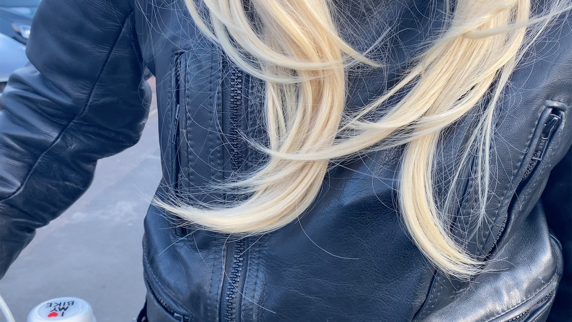 Image of a person in a long blonde wig in motorbike leathers, holding a pushbike with a bell. The design on the bell says 'I love my bike'.  