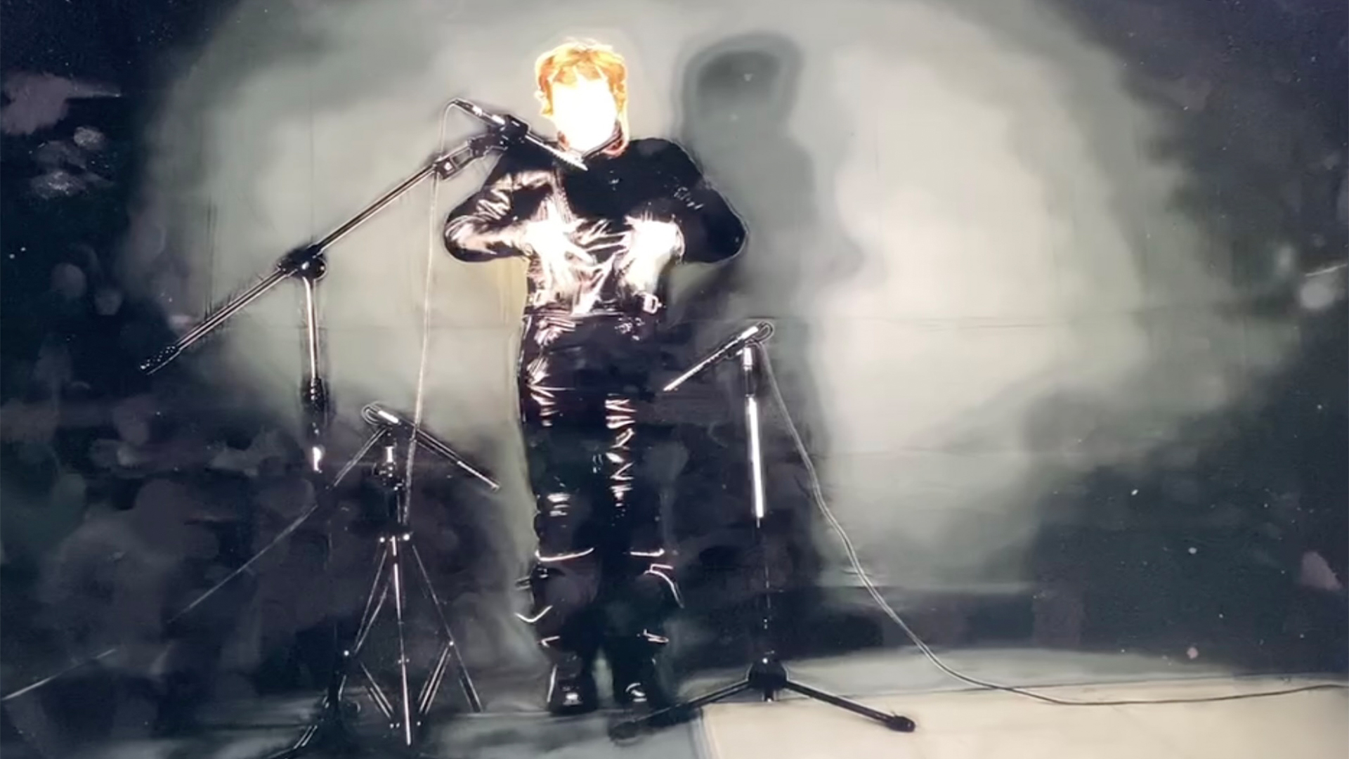 An over-exposed video still of a person in motorbike leathers with their hands rubbing their chest. 