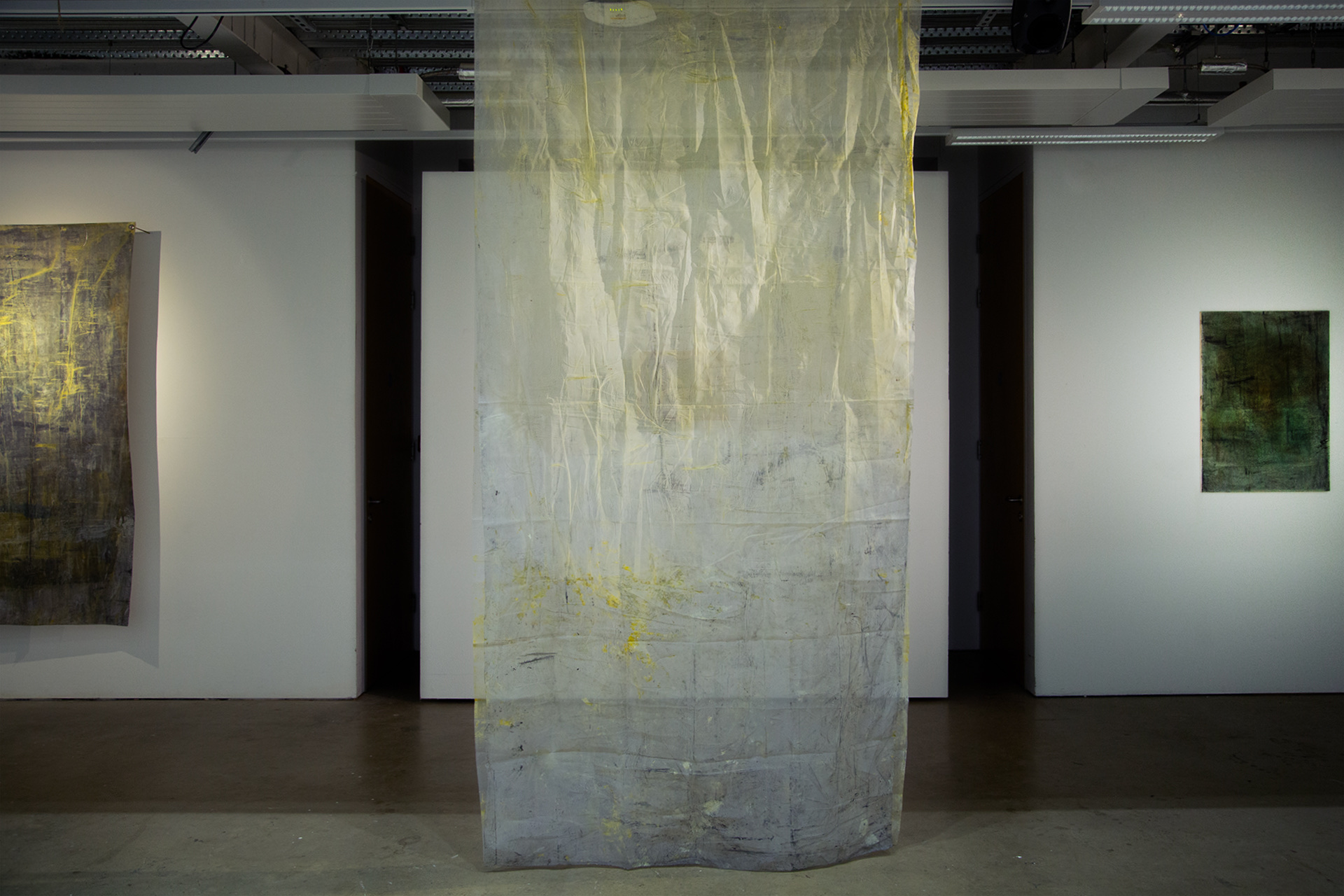 Image of an installation with two abstract green paintings on the back wall and a hanging yellow fabric in the centre of the horizontal image. The fabric is the focus of the image and hangs from the ceiling and touches the floor.