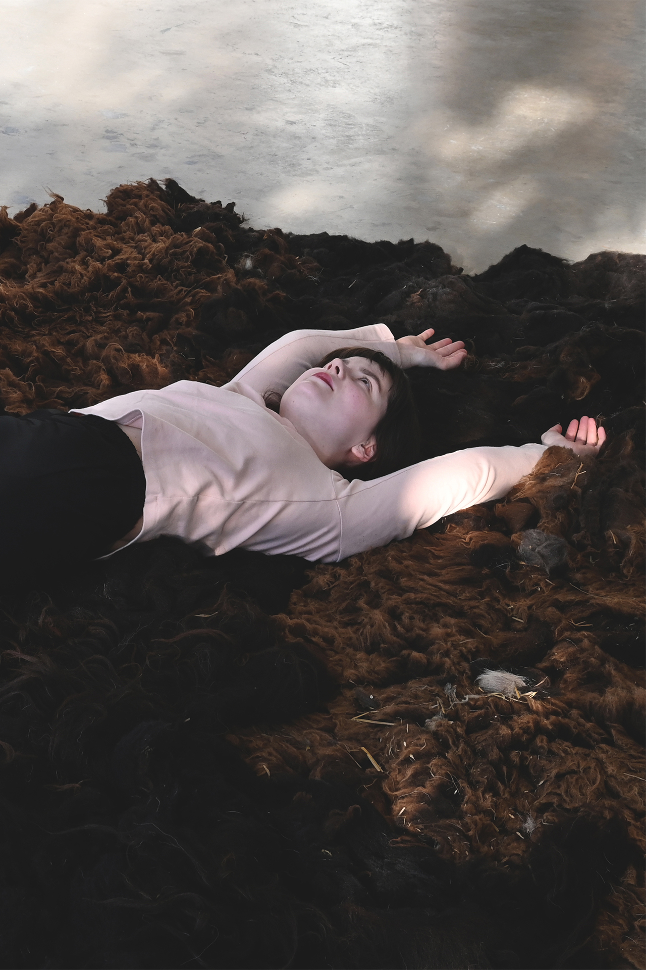 A large amount of brown sheep wool is laid out on the floor. A young woman is lying on the wool, with her arms stretched above her head. The woman is wearing a light pink shirt and black trousers.