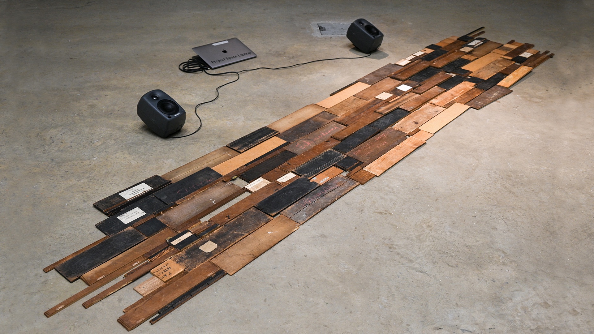 An installation of multiple wooden planks dismantled from lantern slide boxes and arranged on the ground with two Genelec speakers on one side.