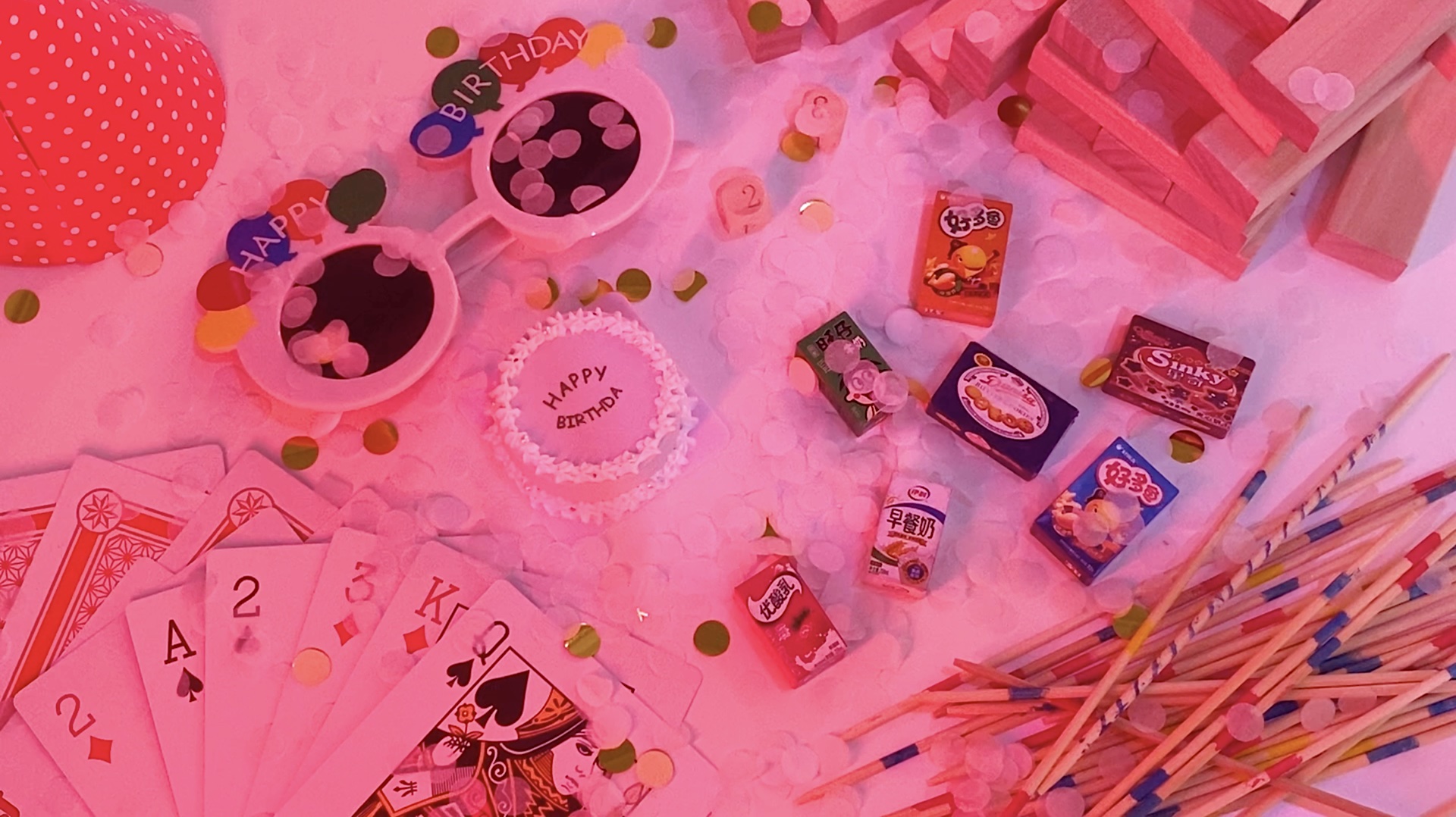 Birthday table set with confetti, a fake birthday cake, playing cards and fake foods and snacks. Shone under fluorescent light.