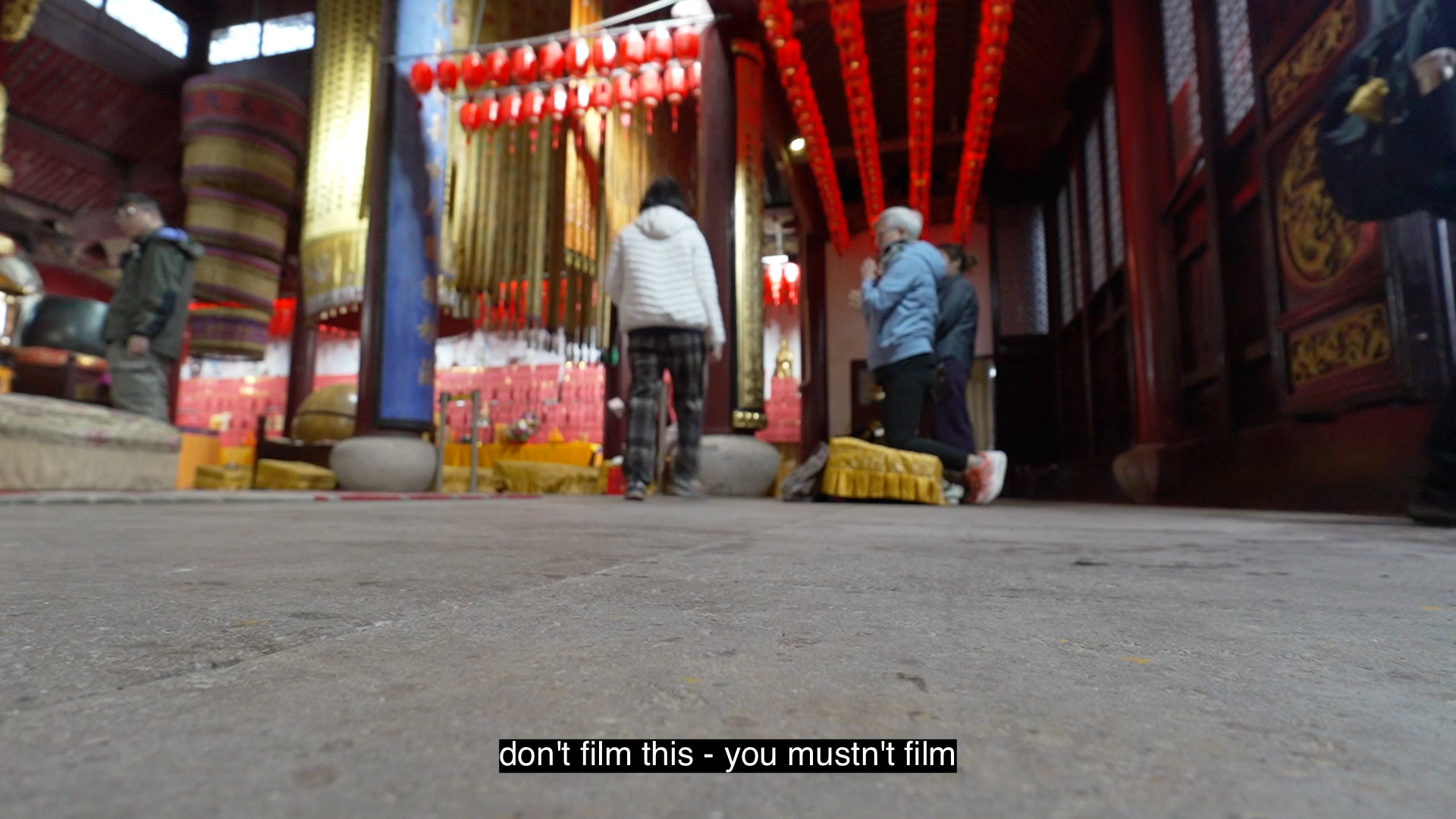A blurred image of the interior of a Buddhist temple, adorned with red lanterns and two people kneeling to pray (captioned: don't film this - you mustn't film)
