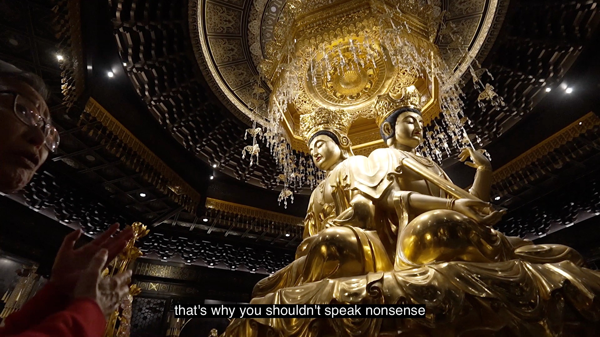 A gold Buddhist statue with two faces, underneath a gold chandelier, with a person speaking to the camera from the left side. (captioned: that's why you shouldn't speak nonsense)