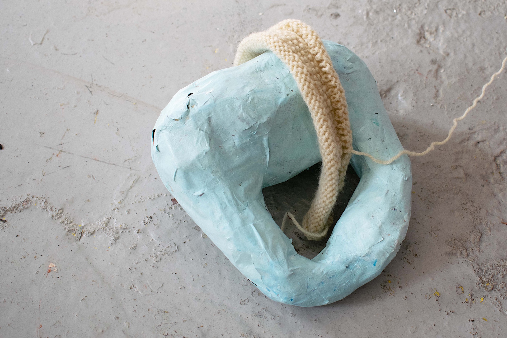 A small pale blue papier mâché sculptural object with a yellow knitted sash, on a grey floor.