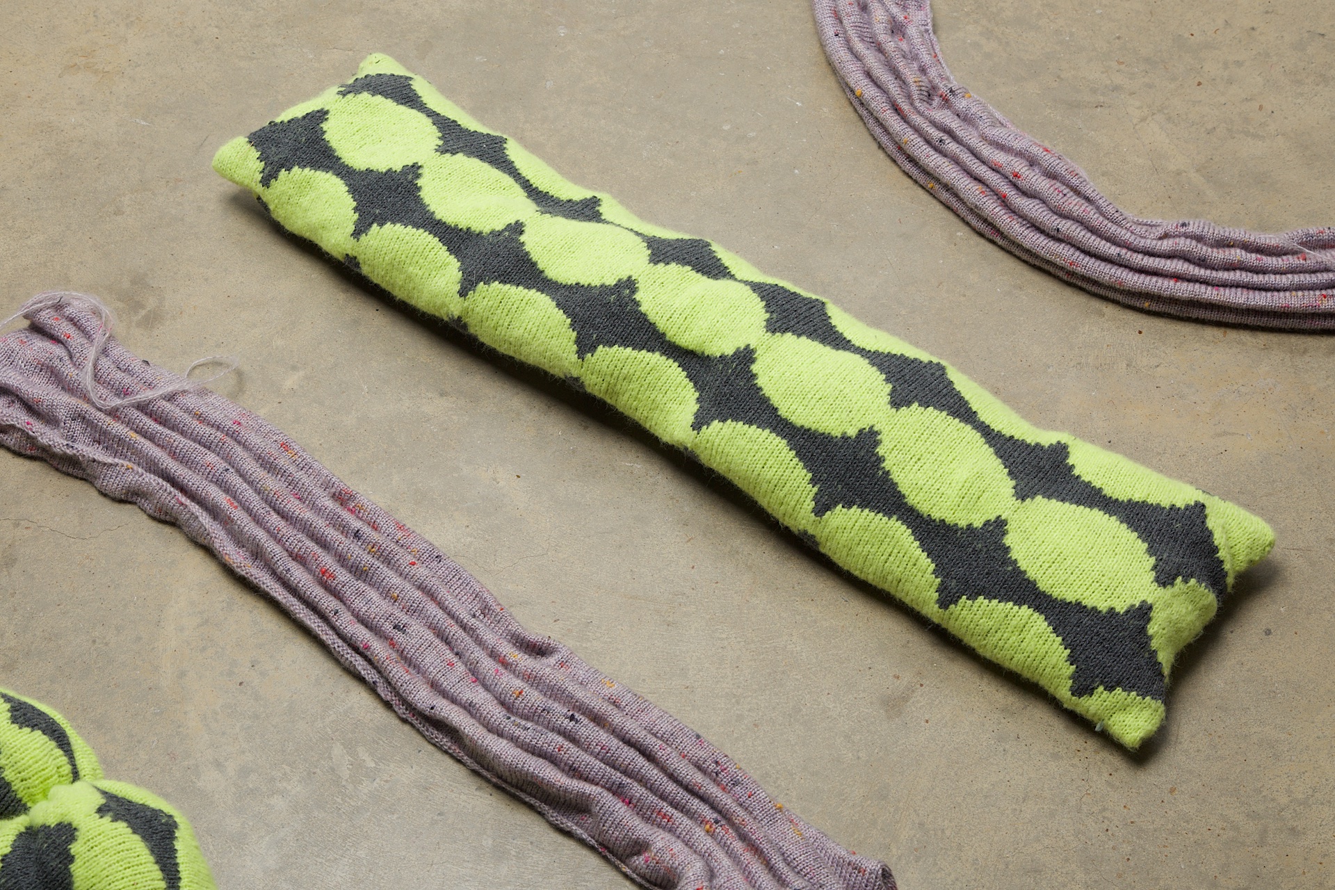 Yellow and grey patterned and purple textured knitted sculptures laying on a grey floor.