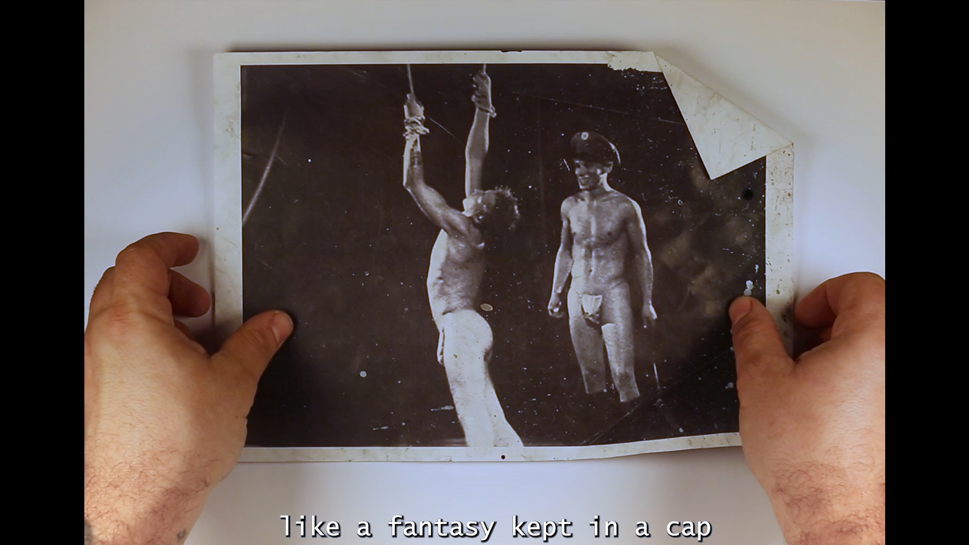 A still from a video, the image shows two hands on a piece of paper. The image on the paper is also a still from a video. The still shows two men engaged in BDSM related to Nazism. A caption at the bottom of the image reads 