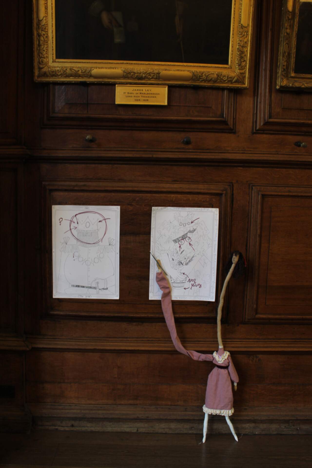 Doll with a long neck in a pink dress holds a paintbrush to two drawings on a wood-panelled wall.