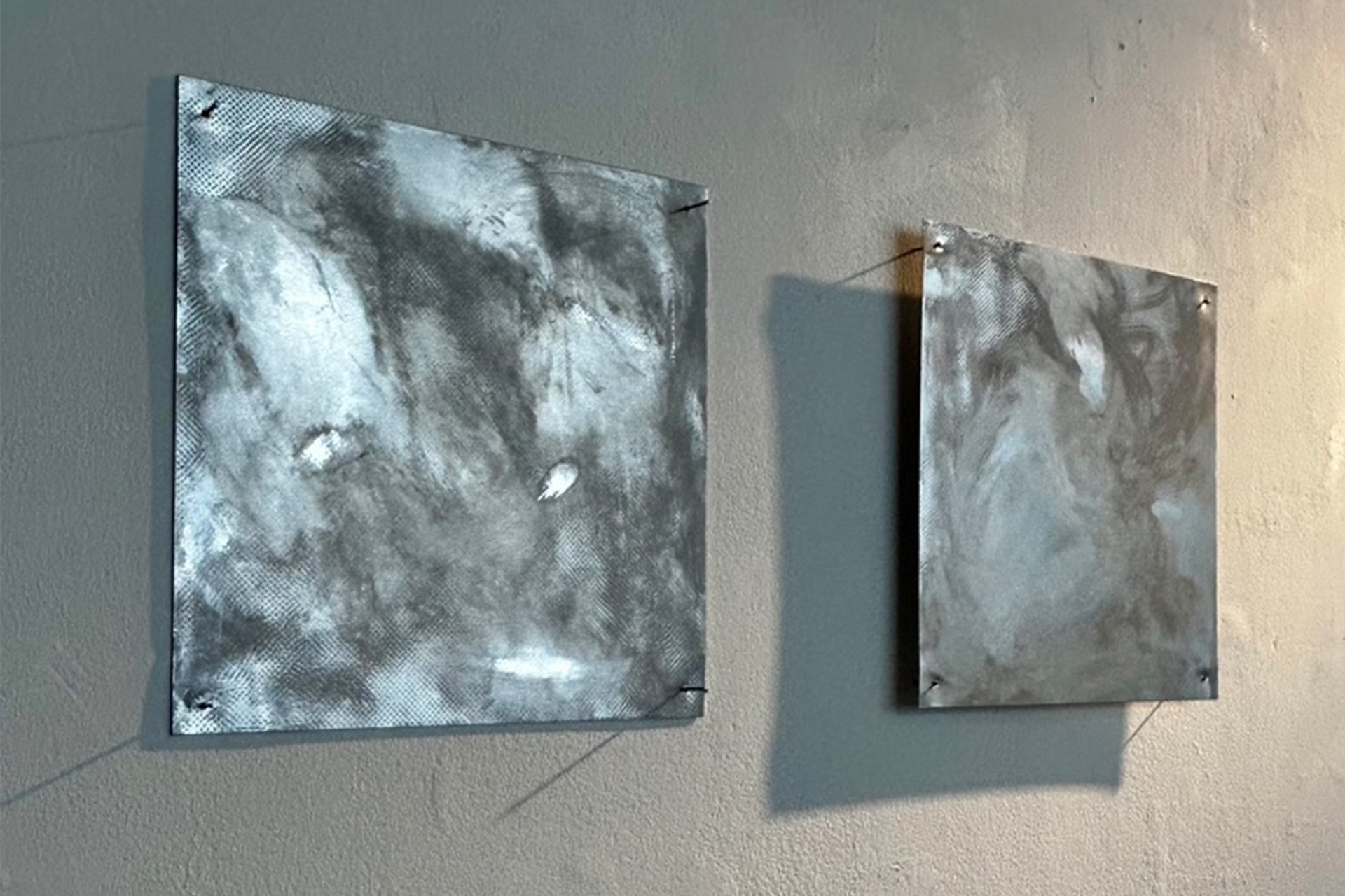 two square sheets of aluminium with blurred, smudged image printed on with transparent medium. There are harsh spotlights lighting the work causing some areas of reflection and warmth where the image is not printed and the mail shines through. The aluminiu