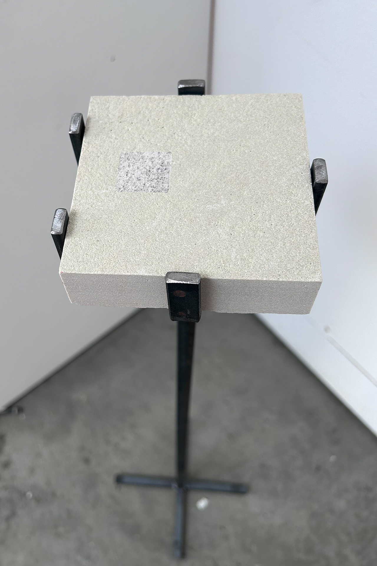 a 100mm square piece of cotswold stone sits inside a metal stand. The stone has a small, square area engraved. The engraving is a blurred, unfocused image that reveals the under-layer of the stones texture, it lies to the bottom left of the stone. The stee