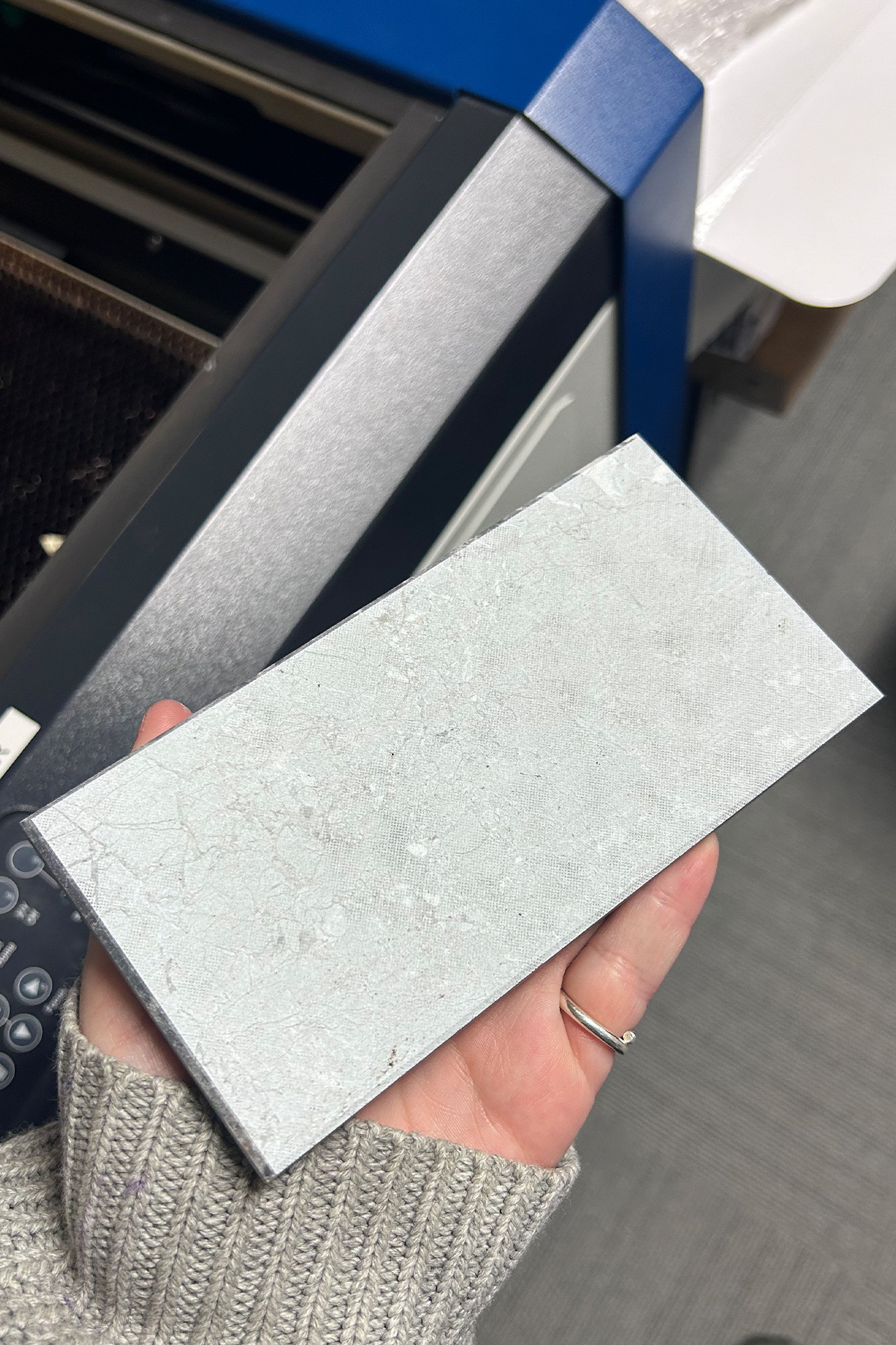 A small rectangular piece of marble laying flat on a hand. The pale grey marble has a small, slight mesh grid engraved onto its surface, this engraving also reveals the deep pattern of the marble swirls. The engraving covers the whole face of the marble sl