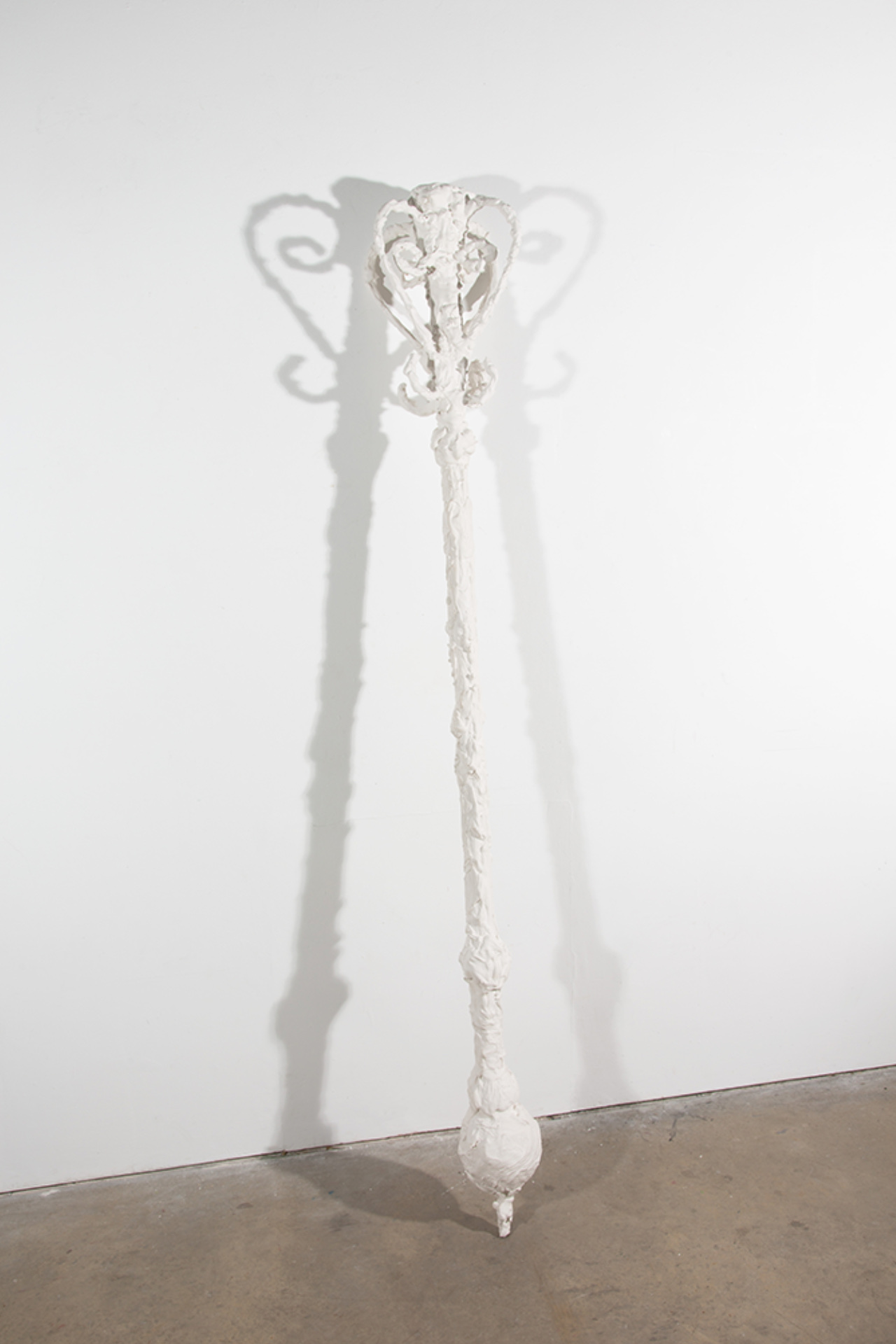 A white sceptre which is made from plaster giving it a texture similar to cream. The height is approximately 2 metres. Here it is displayed leant against a white wall. 