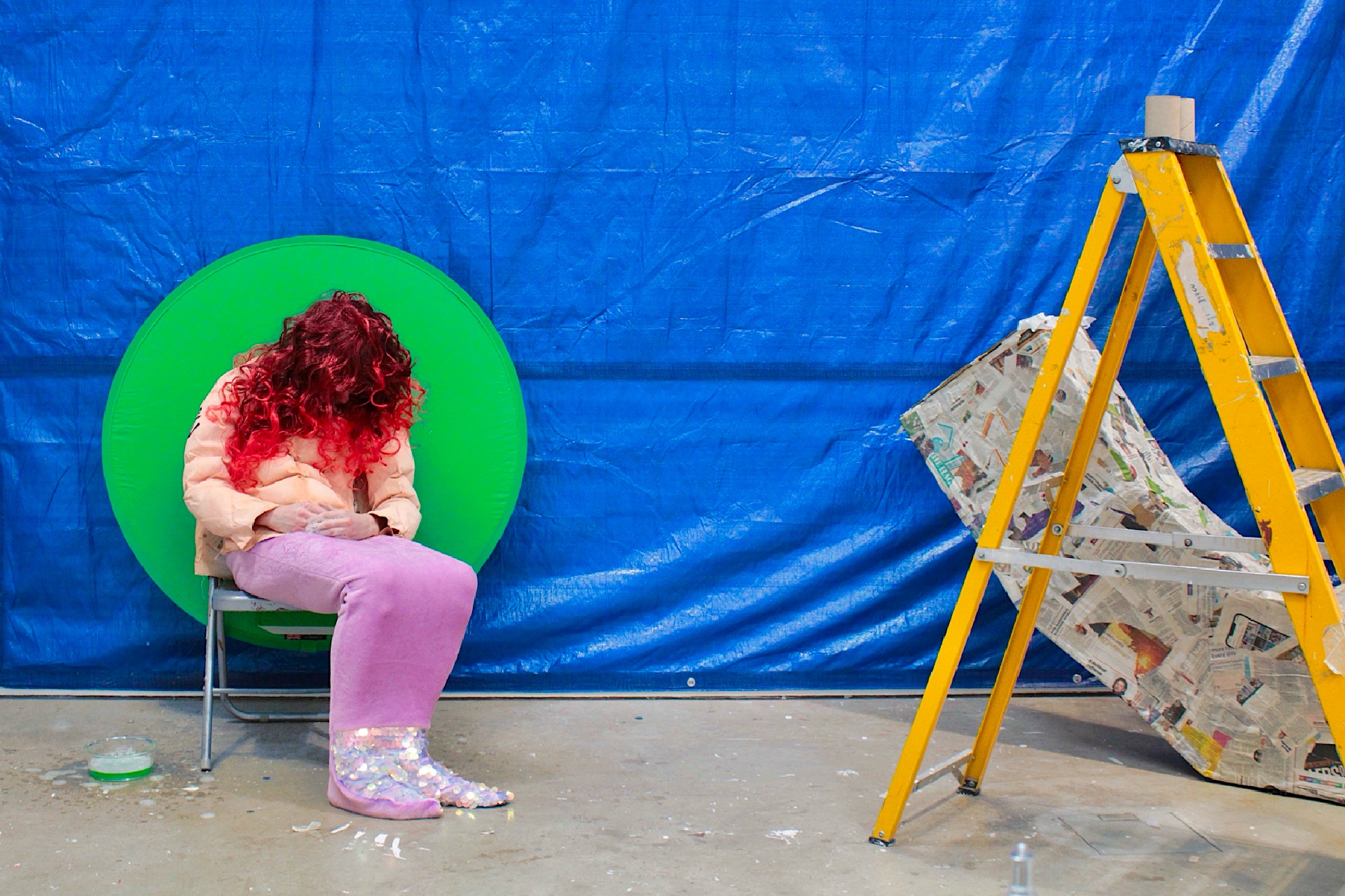 Invigilator figure with a bright red wig, mermaid tail blanket and muscle suit sat on a chair in an installation of a blue backdrop, ladder, cardboard anchor, and bowl of fairy liquid.