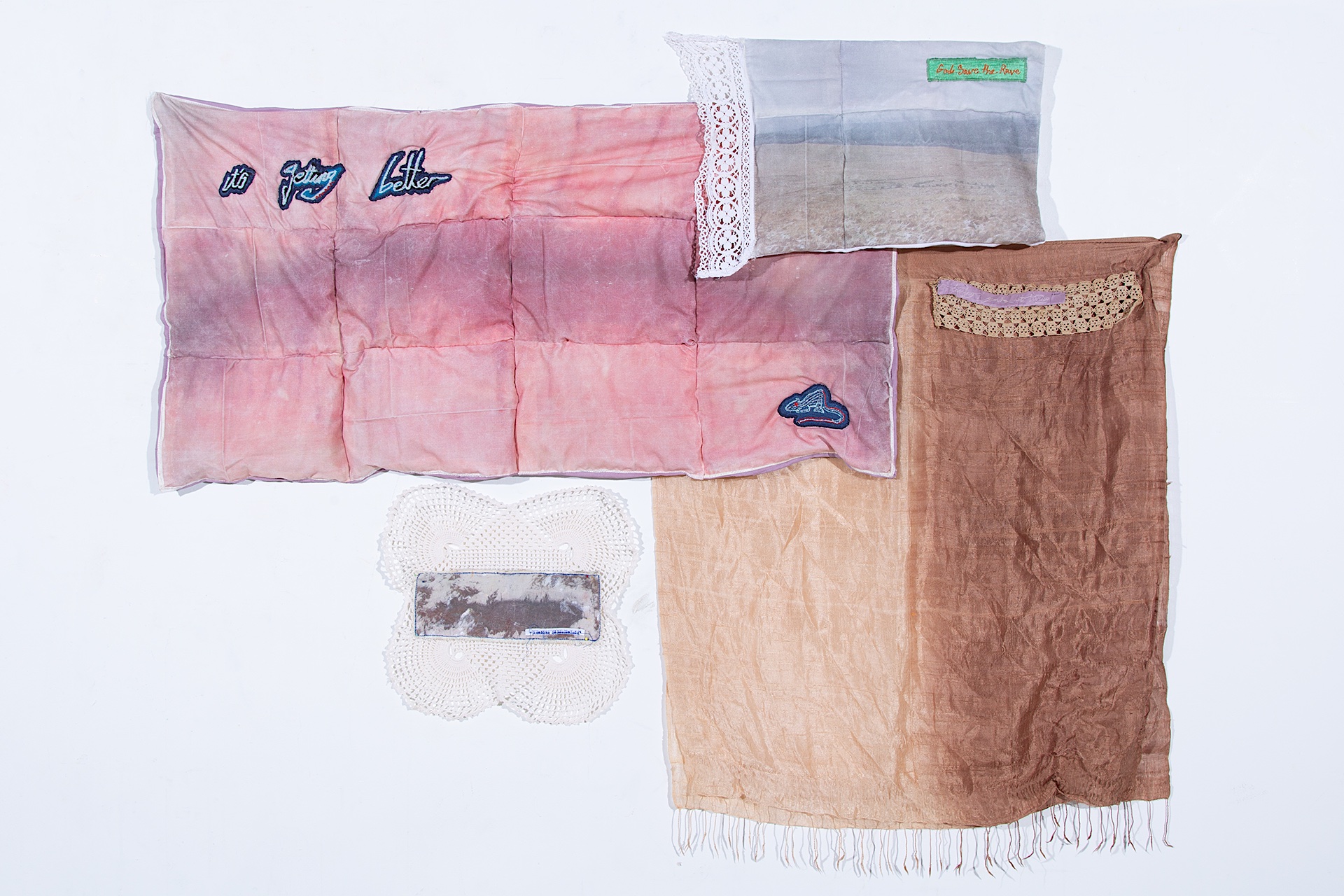 Collection of four textile pieces. TL: pink quilt with embroidered text ‘it’s getting better’ with rat patch. TR quilt with print of a stone circle and embroidered text ‘god save the rave. BL: crochet square with stone circle print. BR: brown silk scarf wi