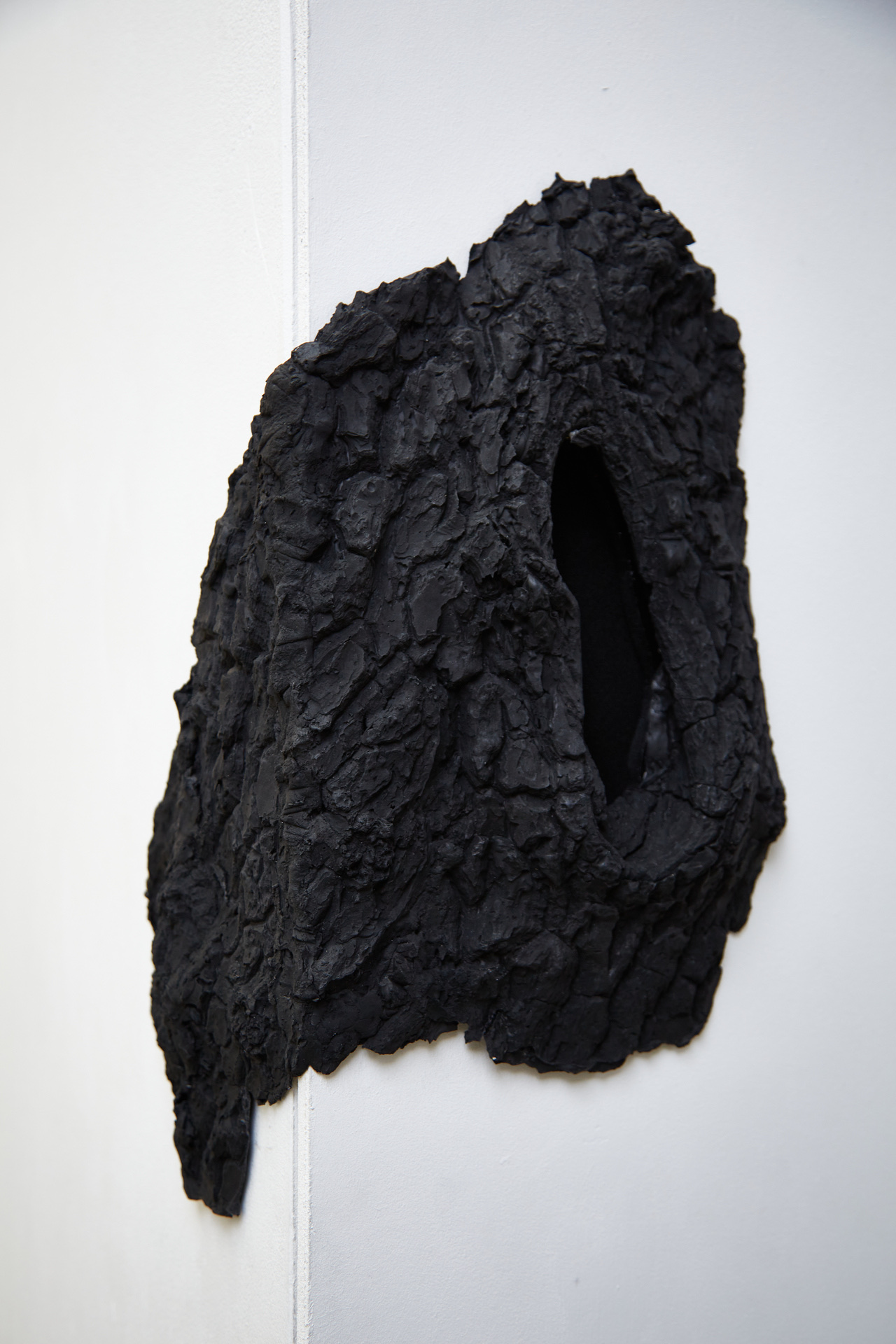 A roughly oval-shaped sculpture, resembling the rugged bark of a charred tree, is mounted on the sharp corner edge of a white gallery wall. The sculpture is entirely black, with a textured, crust-like surface. At its centre is an elongated, dark hollow, al