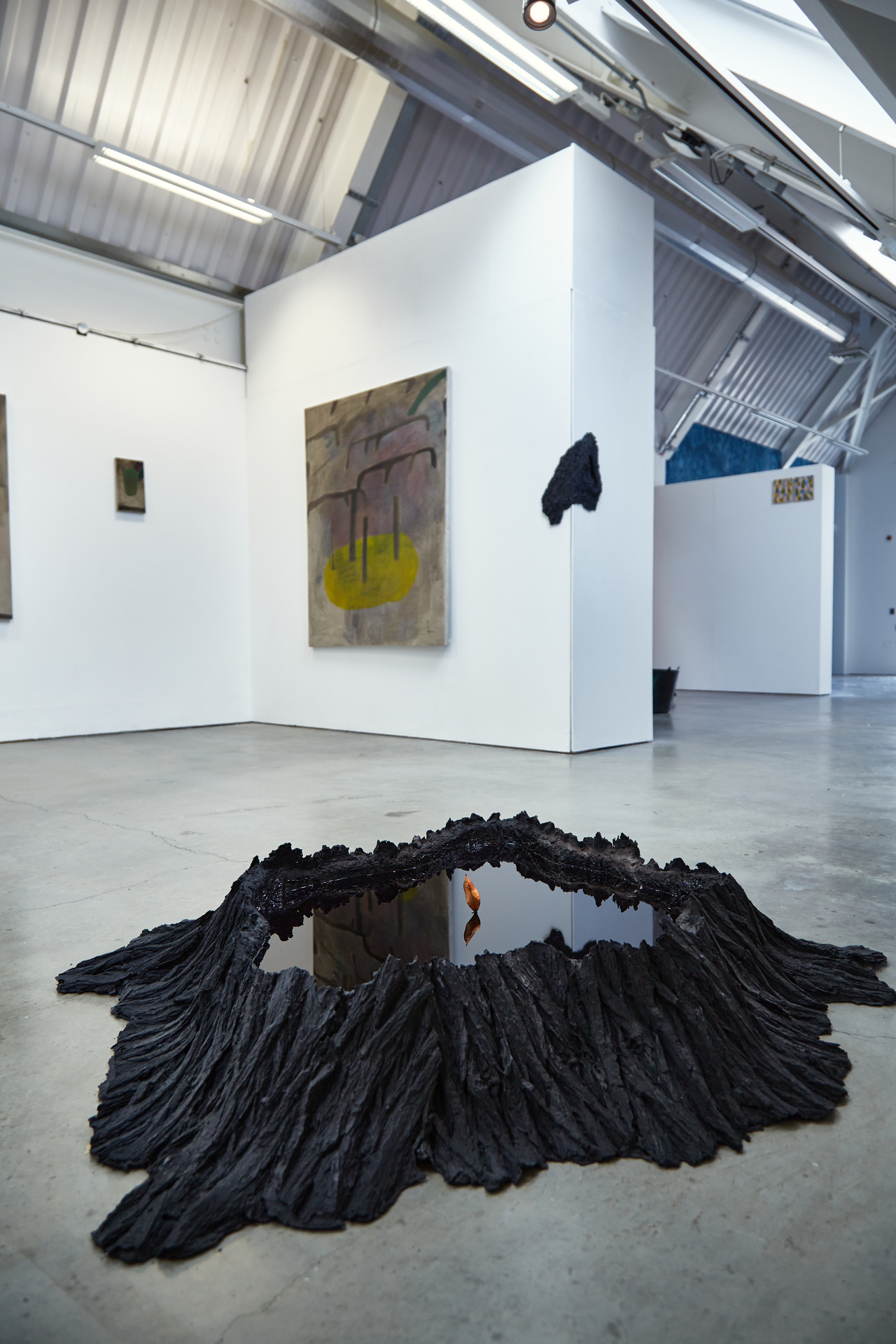 A wide shot of a contemporary gallery space shows a floor installation and wall-mounted sculpture. On the floor, a large, black crater-like form, textured like bark or volcanic rock, contains a smooth black reflective surface, like a pool of still water. A