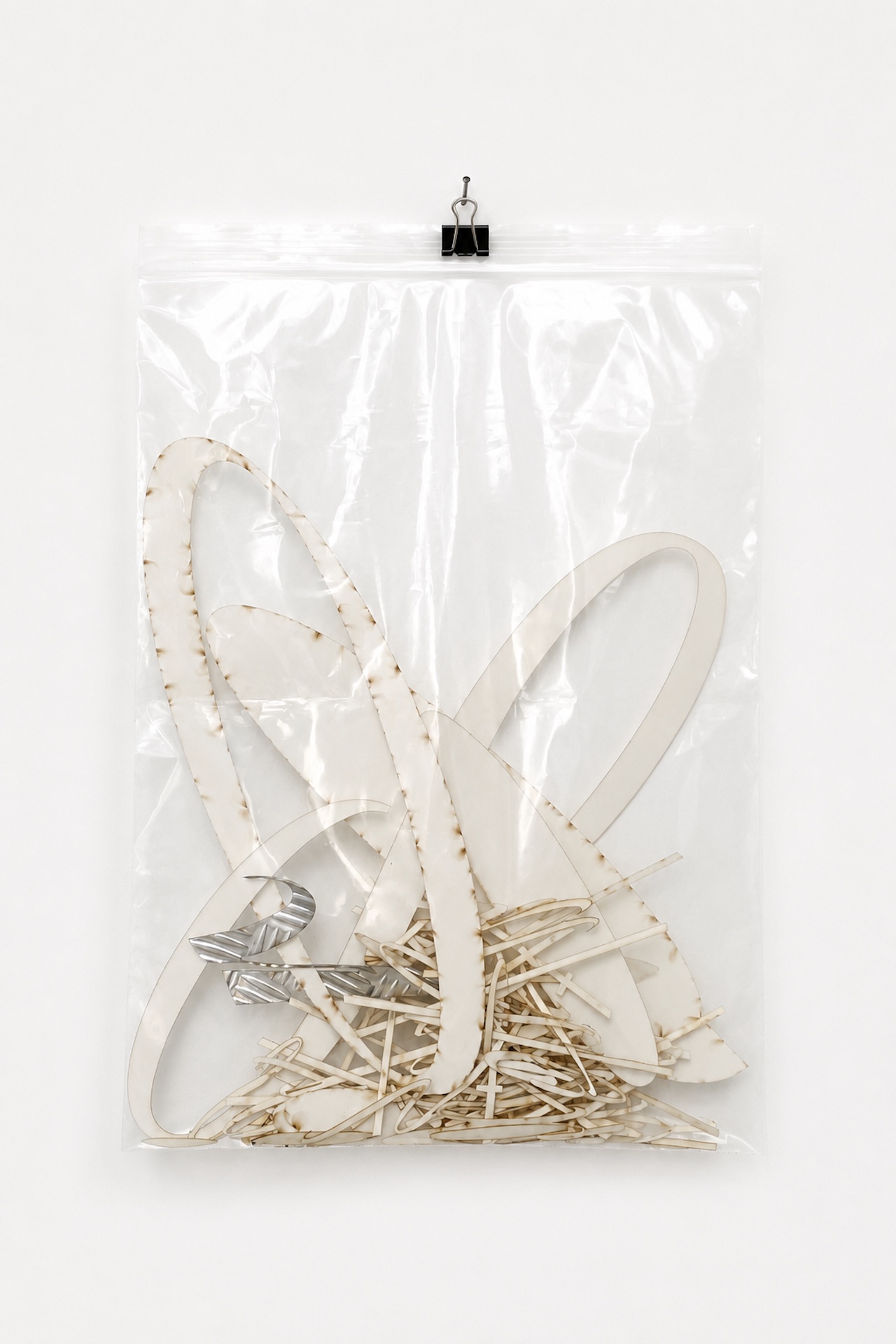 A clear plastic zip-lock pouch containing cream and silver cut out letters hung by a mini black bulldog clip from a nail on a white wall.