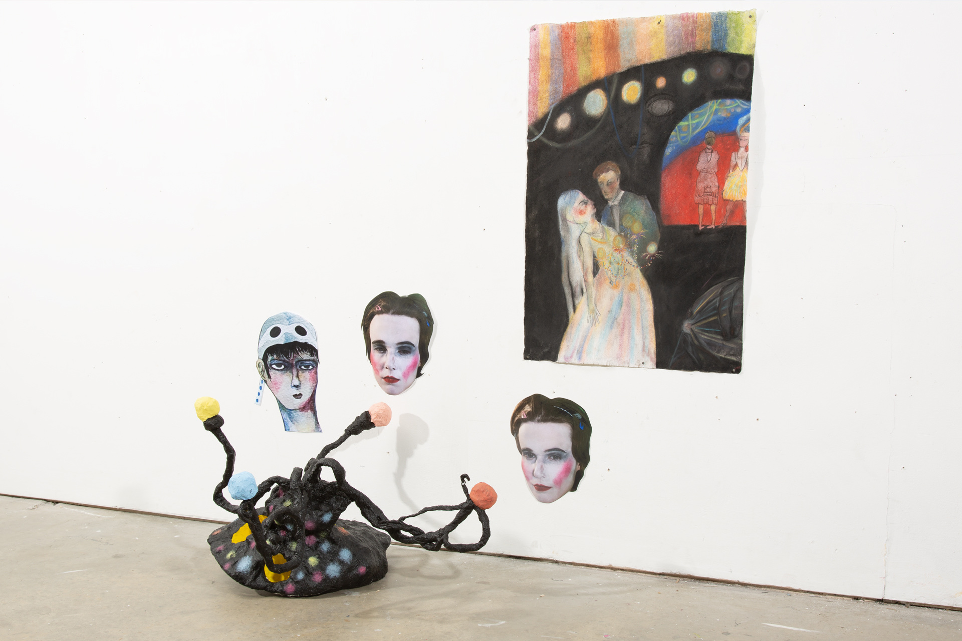 An installation featuring a lo-fi, handmade candelabra in black with blobs of bright yellow, pink and blue on the floor. Beside three of the arms, on the wall, is fixed a cutout head on paper- two photographs of the artist in facepaint, and one a scanned w