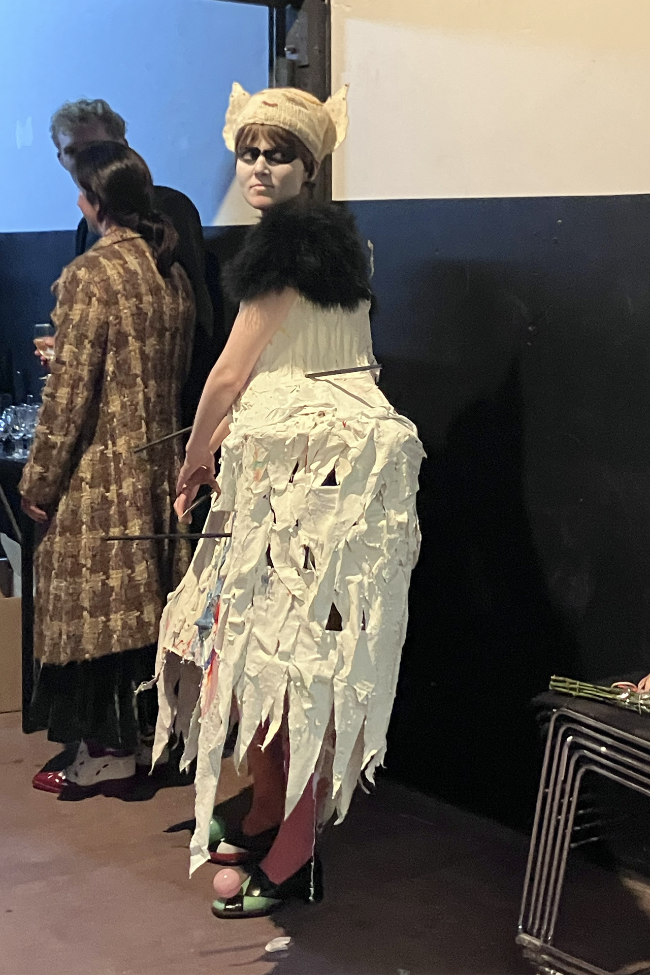 A candid picture. The artist wears a boxy, sculptural dress inspired by the robe-de-style dresses of the 1920s, and circus tents. The dress is covered in triangles of fabric which build in a way that resembles feathers.  She looks off to the right. To the 