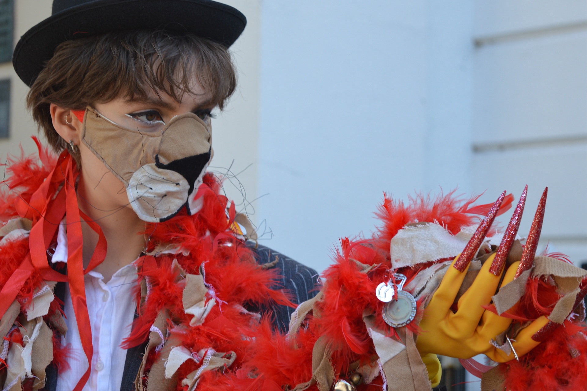 A person with short hair is wearing a lion mask constructed from fabric with wiry whiskers, a bowler hat, a red feather boa embellished with tan fabric and bottle tops, and rubber gloves with long, sparkly, red nails.