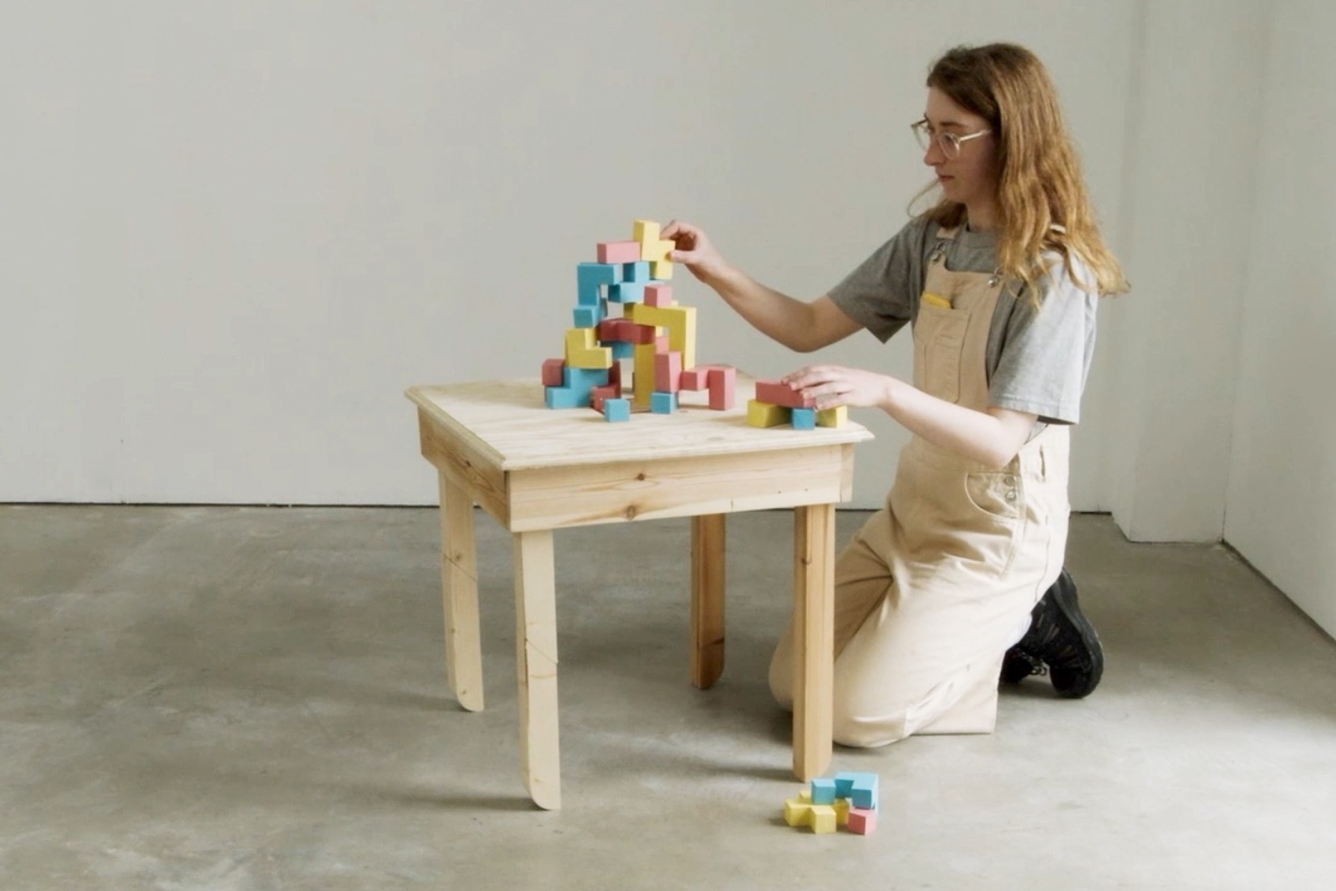 A woman kneels beside a small, wooden table. She builds a tower on top of it out of wooden bricks painted pastel pink, blue and yellow.