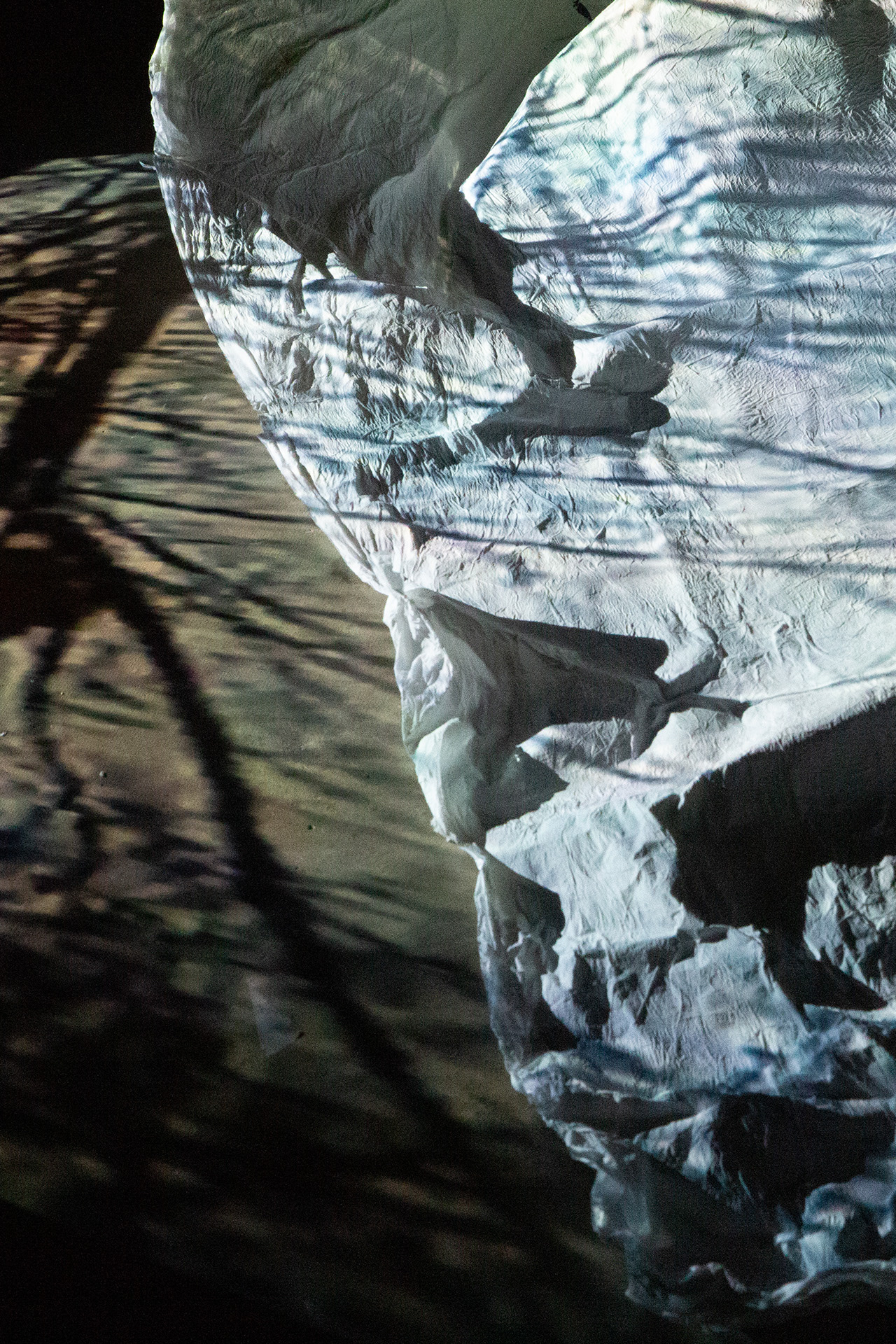 A large piece of creased white paper is gathered on the floor of a dark room. A light projection of tree branches shines across the surface.