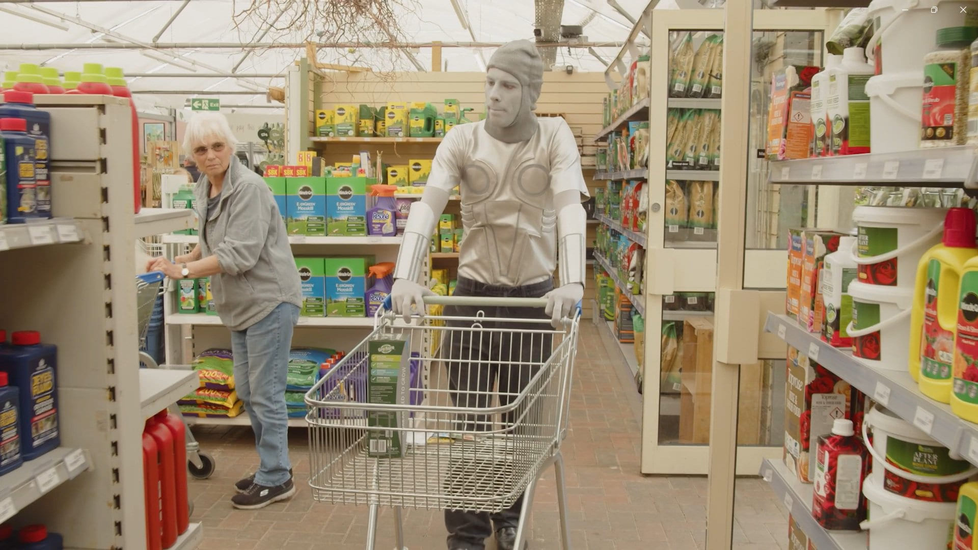 Features a silver figure pushing a shopping cart through a garden centre while an onlooker seems confused 