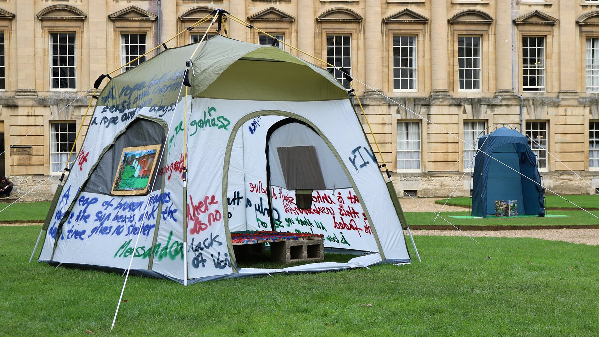 image of two tents. one in the foreground is inscribed with spray painted texts and adorned with paintings on either of its three sides. at the front opening of this tent is a floor mural on a concrete block bed. the tent in the background is a shower tent