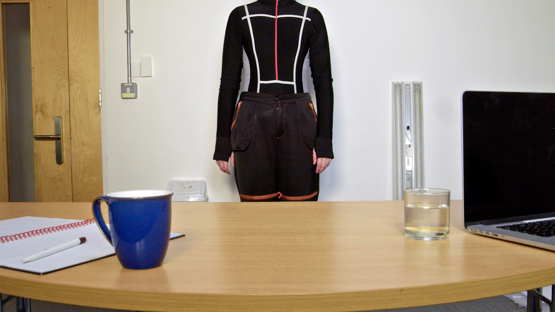 Figure in a black suit with geometric lines, only visible from the neck down, stood behind a desk.