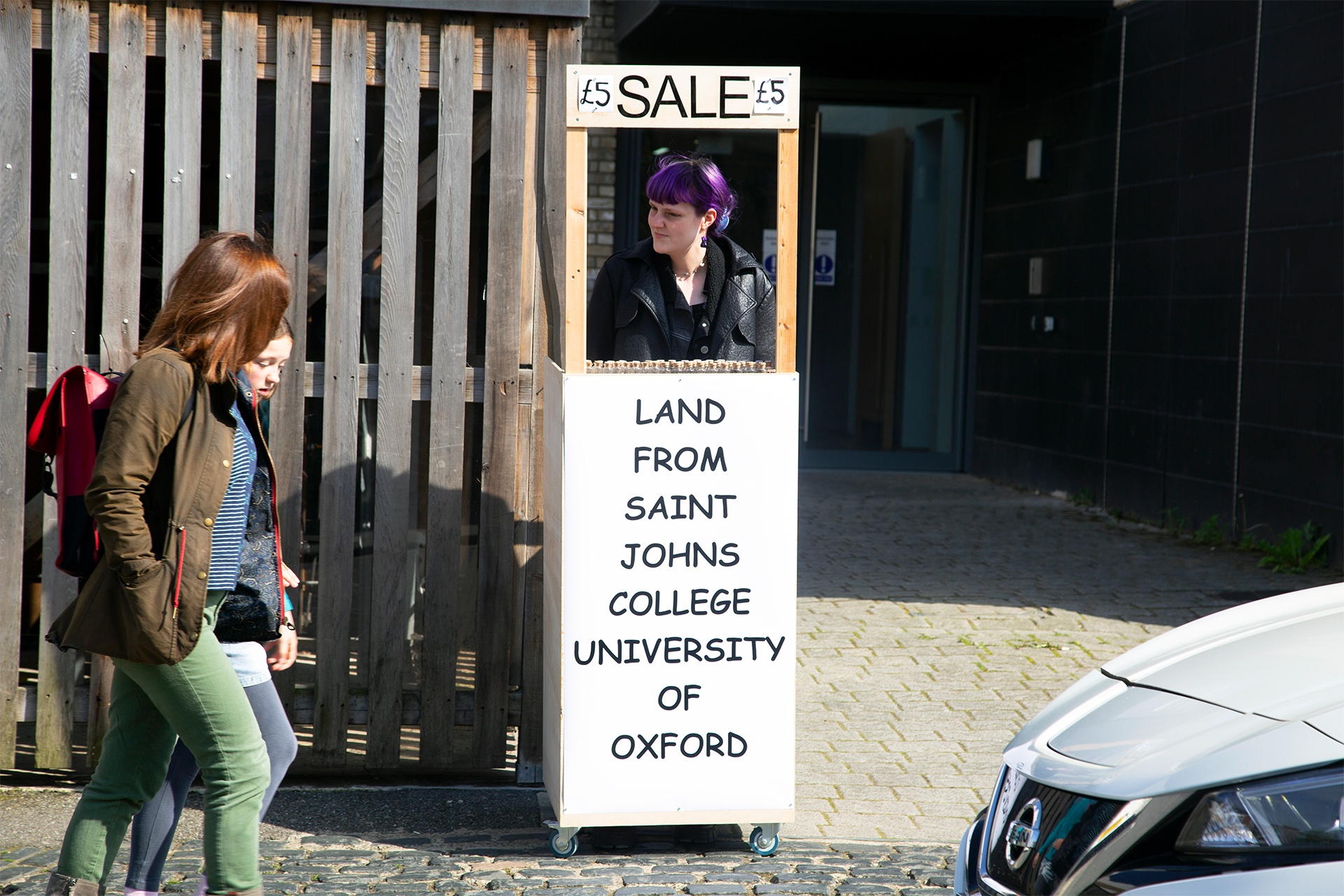 Figure on busy street with stall reading ‘SALE: Land from Saint Johns College University of Oxford’