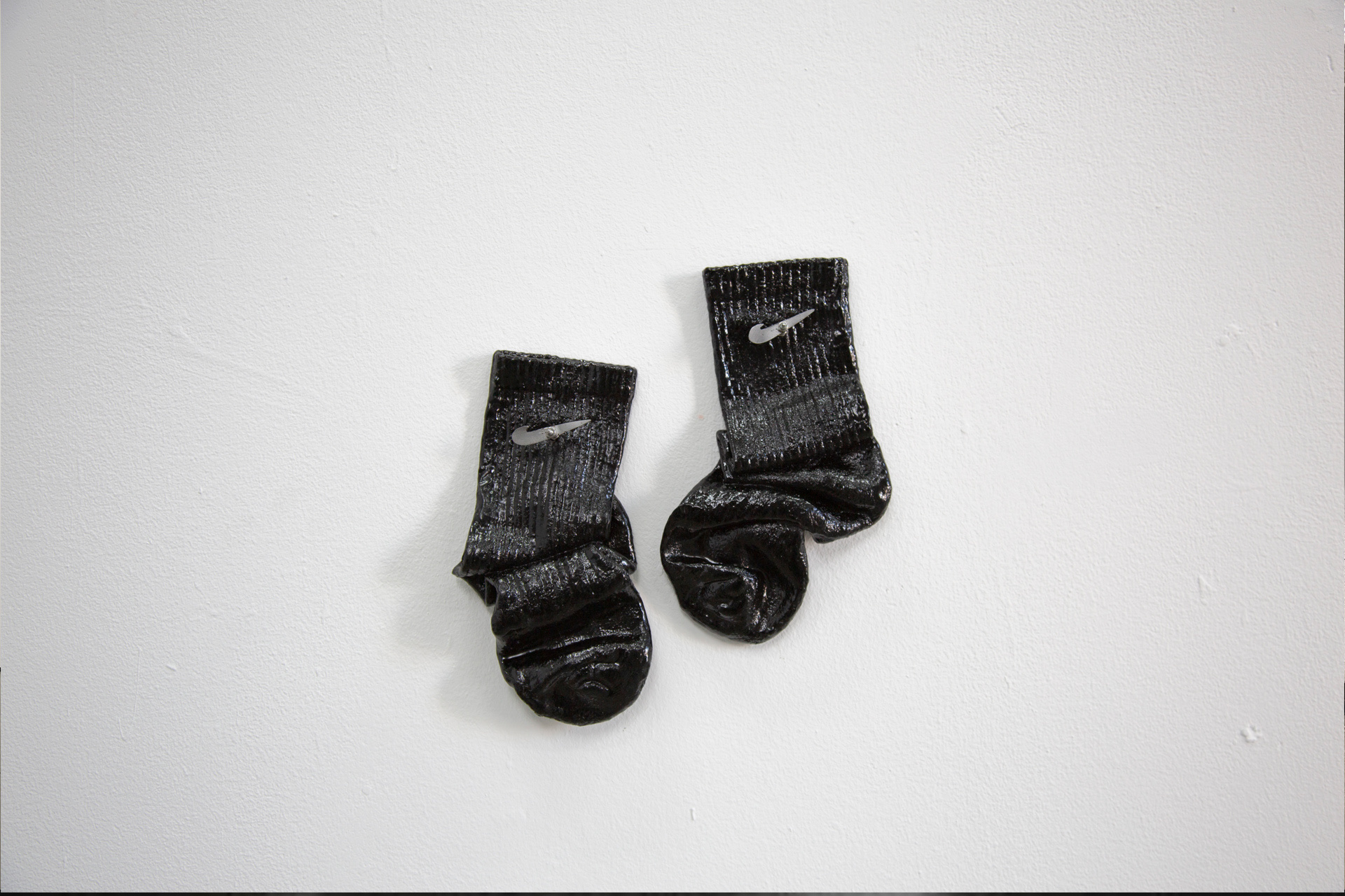 A sculpture depicting a pair of black socks, crumpled as if discarded after a long day. The socks are soaked in tar to create a firm and sticky-looking object. Sharp metal tick motifs protrude from the upper part of the socks.