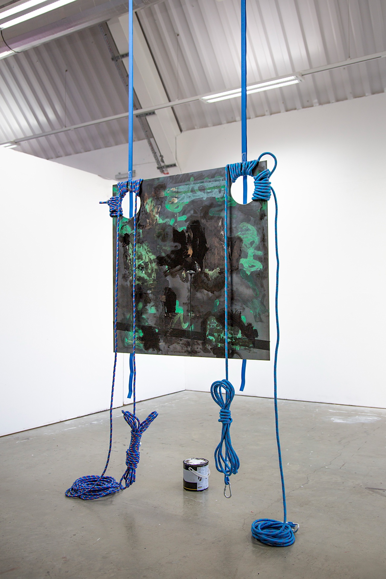 Suspended are two sculptures made from steel. In the foreground, a steel ball, composed of multiple interconnected parts, sits on the floor, suspended from the ceiling by a chain, alongside sharp, polished metal pieces. Towards the back, two metal hands la