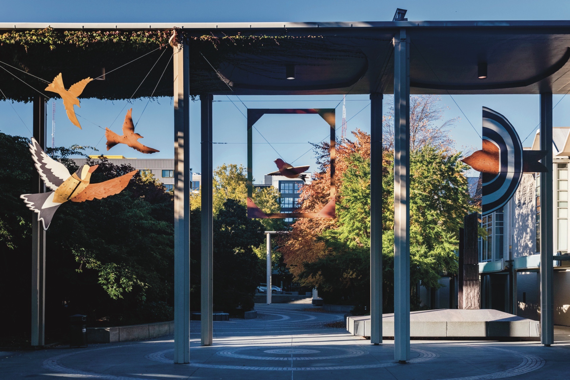 An installation of three bird sculptures hung under a tall covered walkway. The birds are installed in between the columns. The birds are cut out of wood. Some areas are painted and others are stained wood. In the middle an 8 x 10 ft frame is hung with one