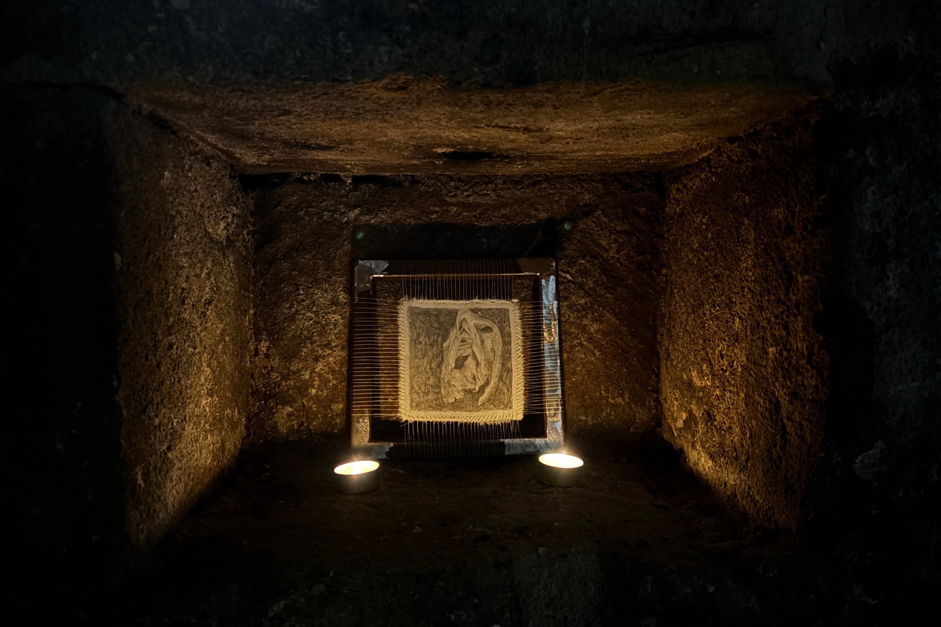 Image shows a drawing of seaweed stretched on fabric on a metal frame which is candlelight in a niche in a crypt.