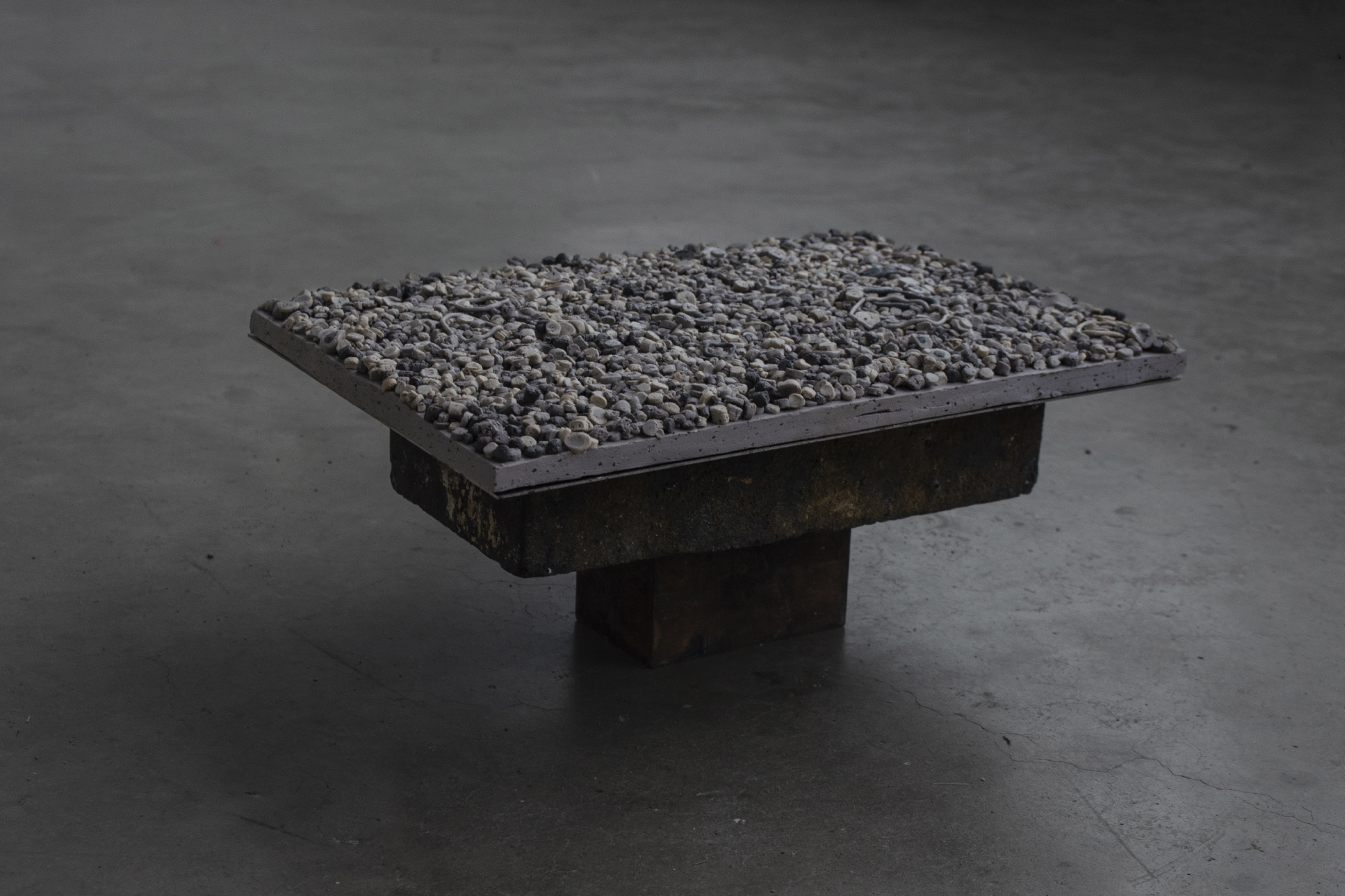 A flat, rectangular light-grey sculpture composed of hundreds of small clay and sand circles, densely arranged to form its surface. It rests on a thin sheet of steel, supported by a low plinth made of brick and a solid block of steel.