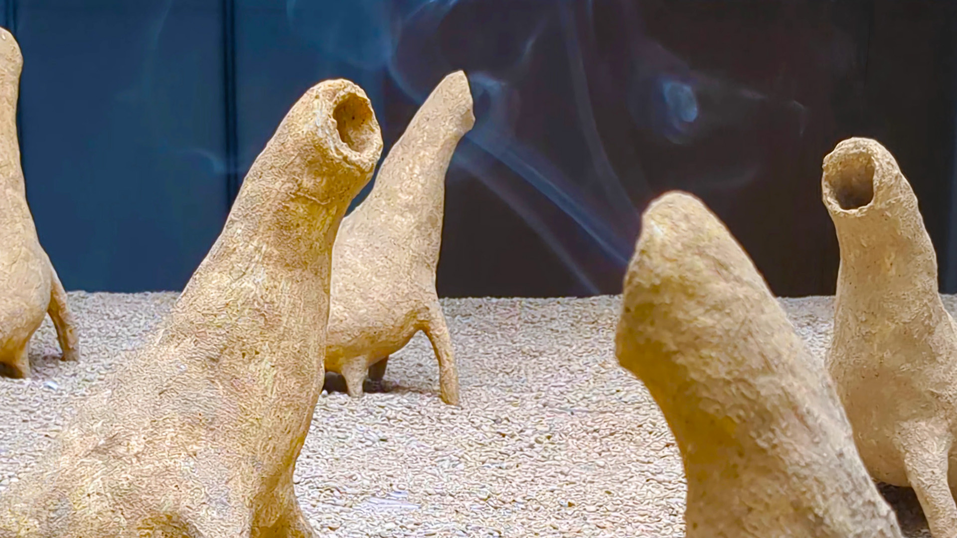 Five creatures on a flat sandy horizontal surface hung from the ceiling. The creatures are all belching out smoke from their mouths, facing different directions. They are abstract and resemble dinosaurs. Each of them have a long neck and four legs. The mon