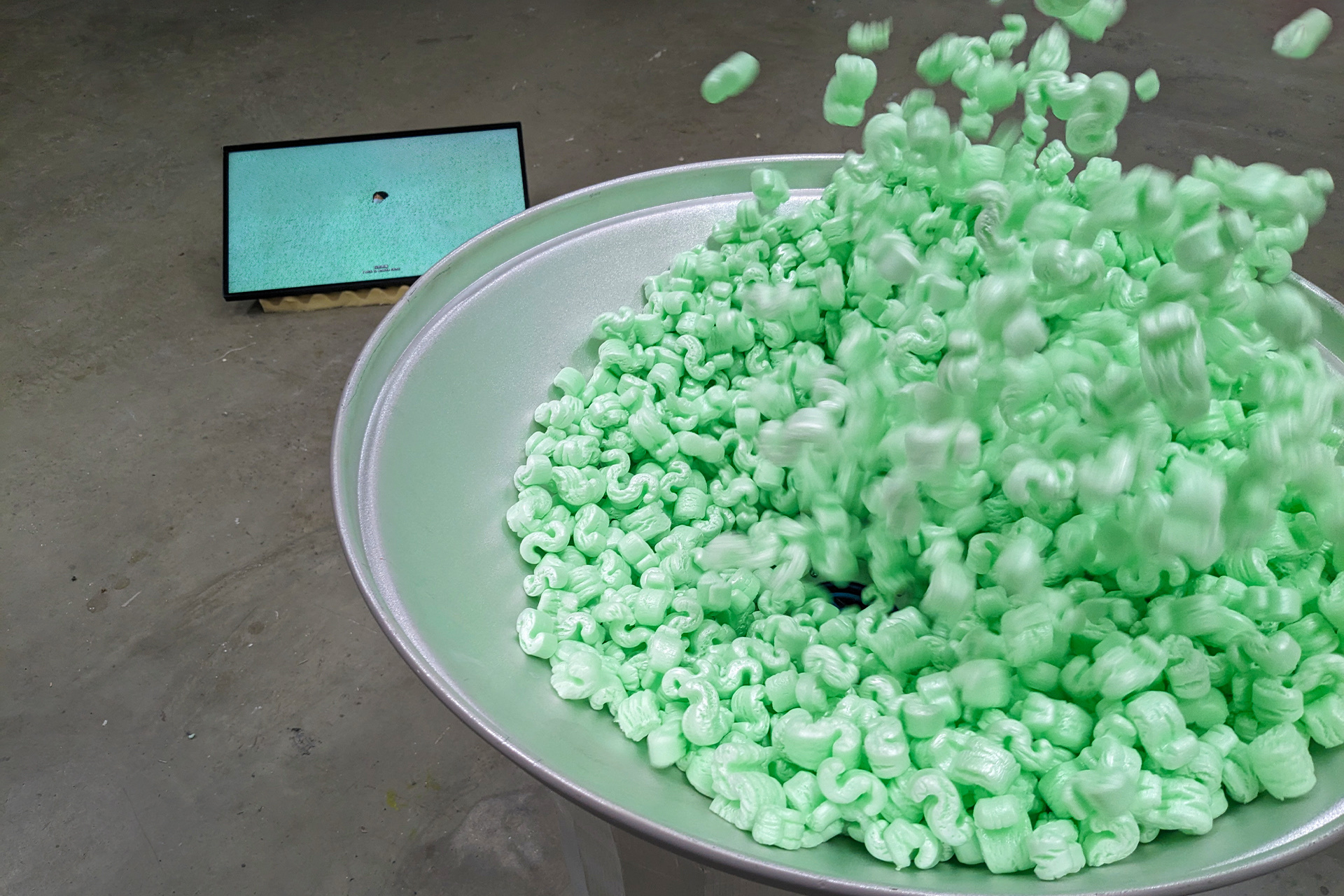 On the left, there is a TV screen on a yellow piece of soundproofing foam on the ground. The screen is playing a video of a person with facial mask, eyes closed, in a sea of green S-shaped packing peanuts. Only the person's head is seen amidst the packing 