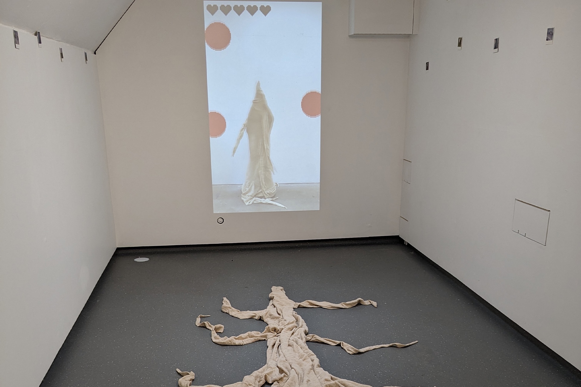 The photo shows the middle wall, long left and right walls and sparkly floor of a narrow gallery space 2.8 metres wide and 7 metres long. On the middle wall of the gallery, a video projection shows a character wearing a cotton dust sheet costume of a nerve